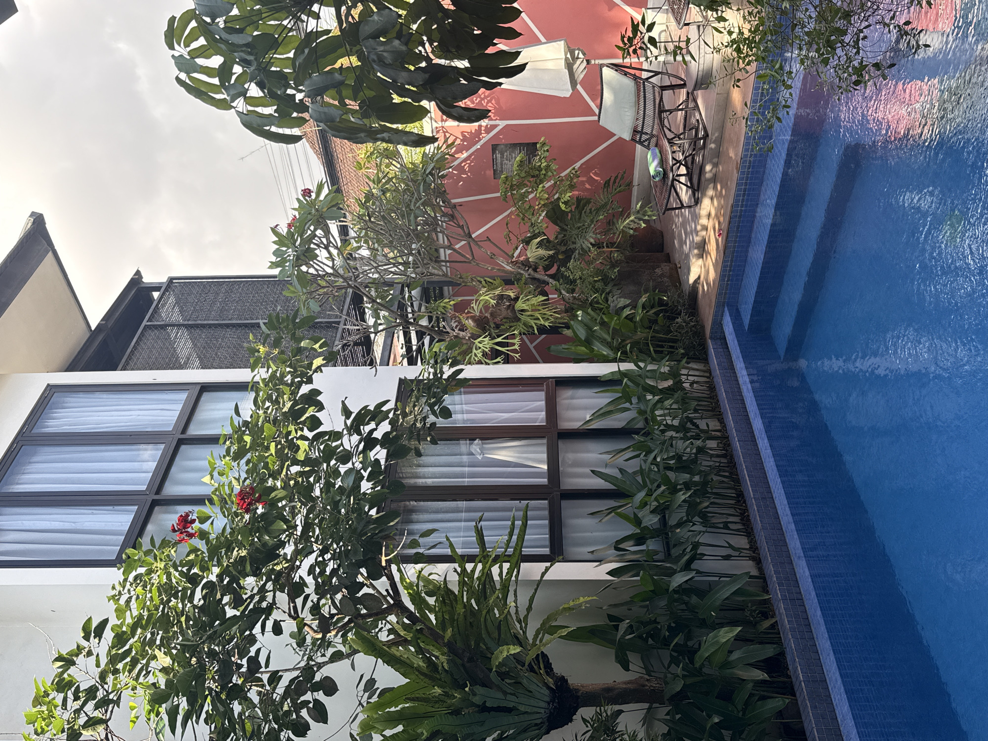 The image captures a view in North Kuta, Indonesia, showcasing a building with a pool area. The building, likely a villa or residence, is painted white and has multiple windows with dark frames. A tree with red flowers grows near the building. Adjacent to the building is a swimming pool with blue tiles lining its edge. The pool water is a bright blue color, reflecting light. In the background, a pink building with white geometric patterns is visible. Garden chairs are placed beside the pink building. Foliage and other plants are prominent throughout the scene. The image is slightly tilted.