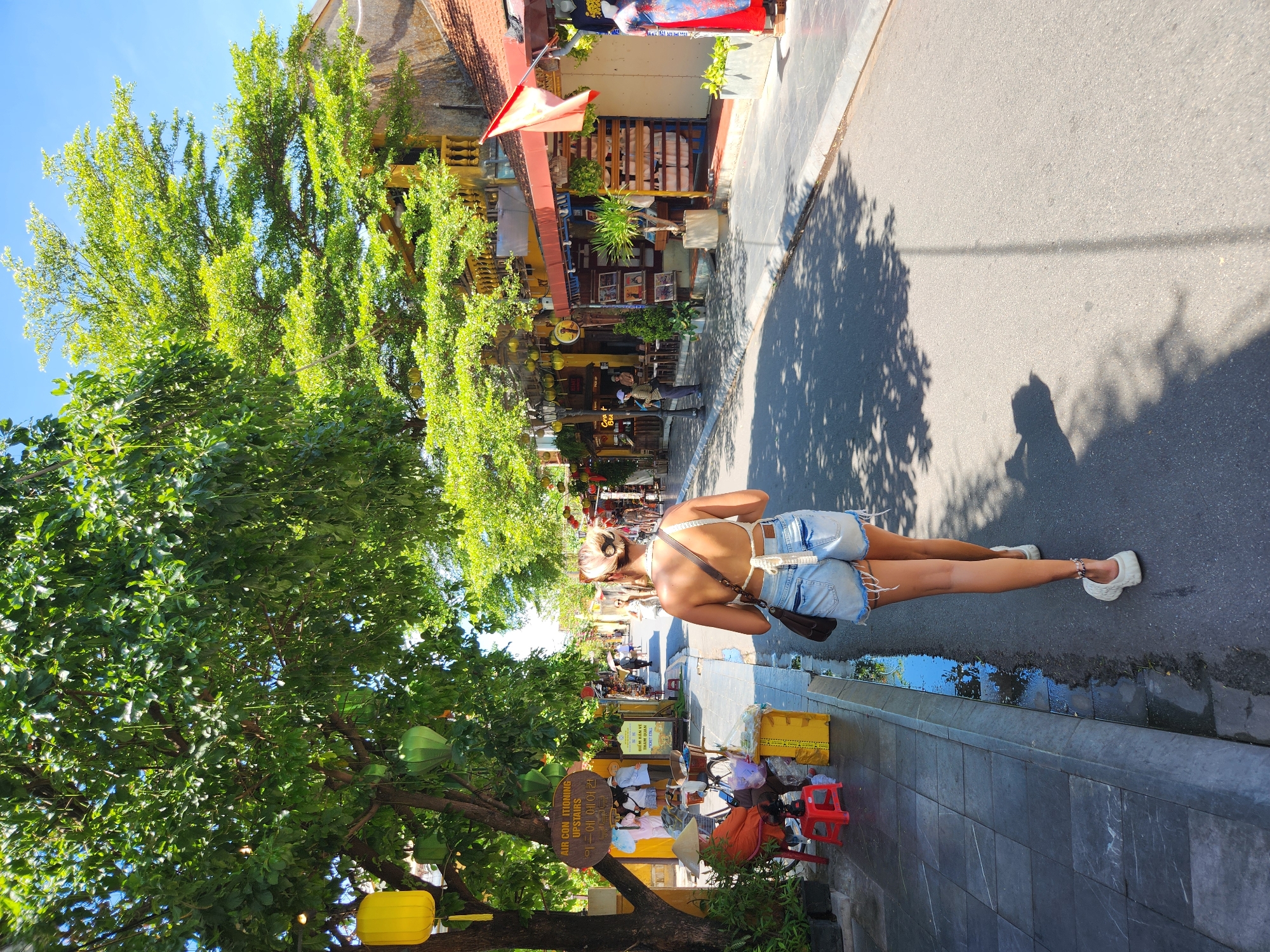 In Hội An Ward, Vietnam, on a sunny day, a young woman with blonde hair pulled back in a bun walks down a paved street. She is wearing a white halter top with a crocheted detail, light-wash denim shorts with frayed hems, and white slip-on shoes. She carries a brown crossbody bag. The street is lined with buildings with yellow facades and balconies, and shops with displays of goods, including clothing and art. Large trees with lush green foliage provide shade over the sidewalk. Yellow and green lanterns hang from the trees. On the sidewalk, a vendor sits on a red stool next to a selection of items, including a yellow cooler and plants. Several other people are visible in the background, some walking and others standing near shops. The street shows signs of recent rain, with a puddle of water reflecting the sky and surrounding trees along the curb. The overall scene depicts a bustling commercial area in a Vietnamese city under a clear blue sky.