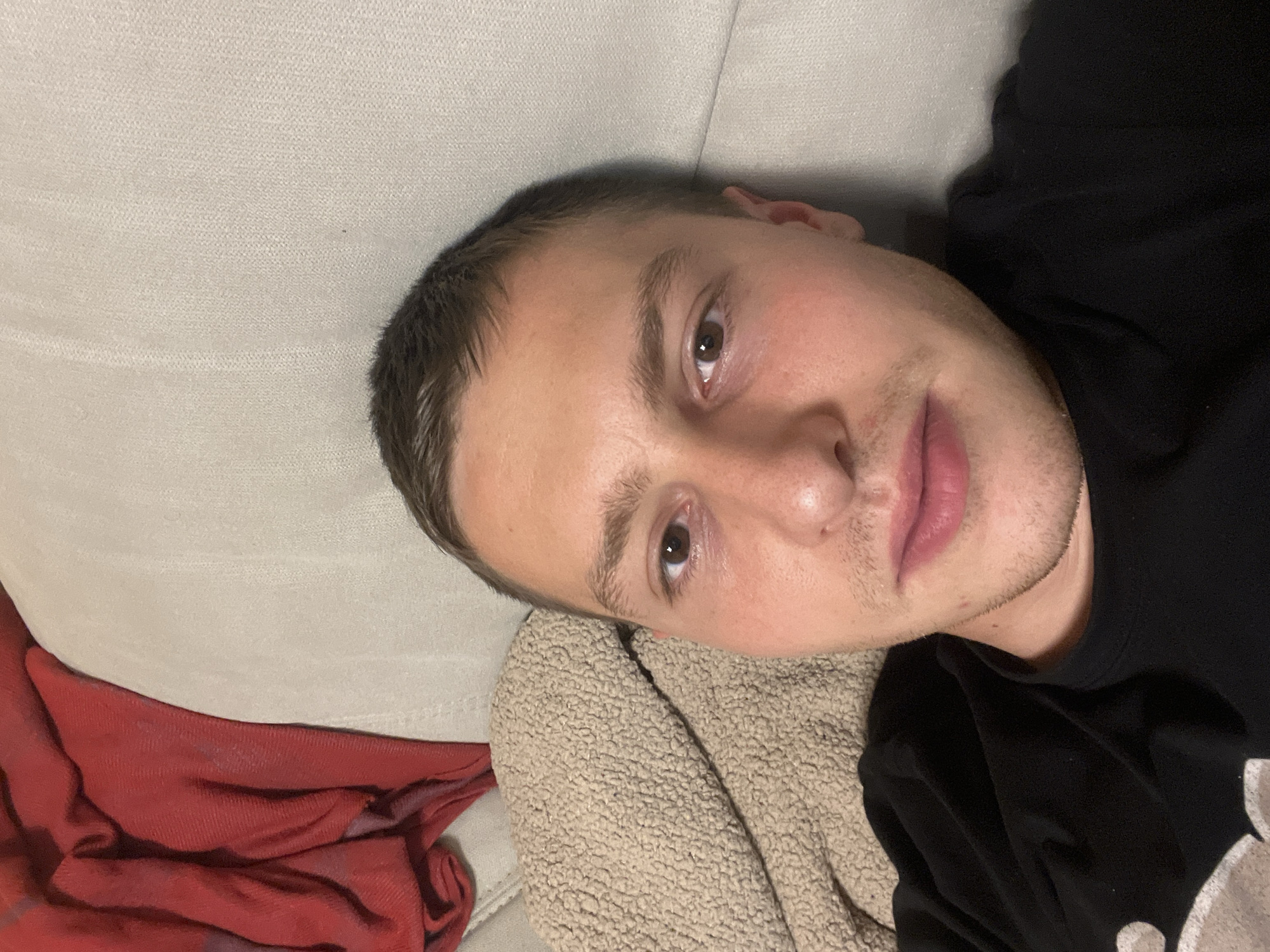 A young man is lying on a couch. He is fair-skinned with short brown hair, brown eyes, and stubble. He wears a black shirt. His head rests on a tan-colored blanket or pillow. A red blanket is visible to the left of the man. The background consists of a light-colored couch. There is no location information provided in the image.