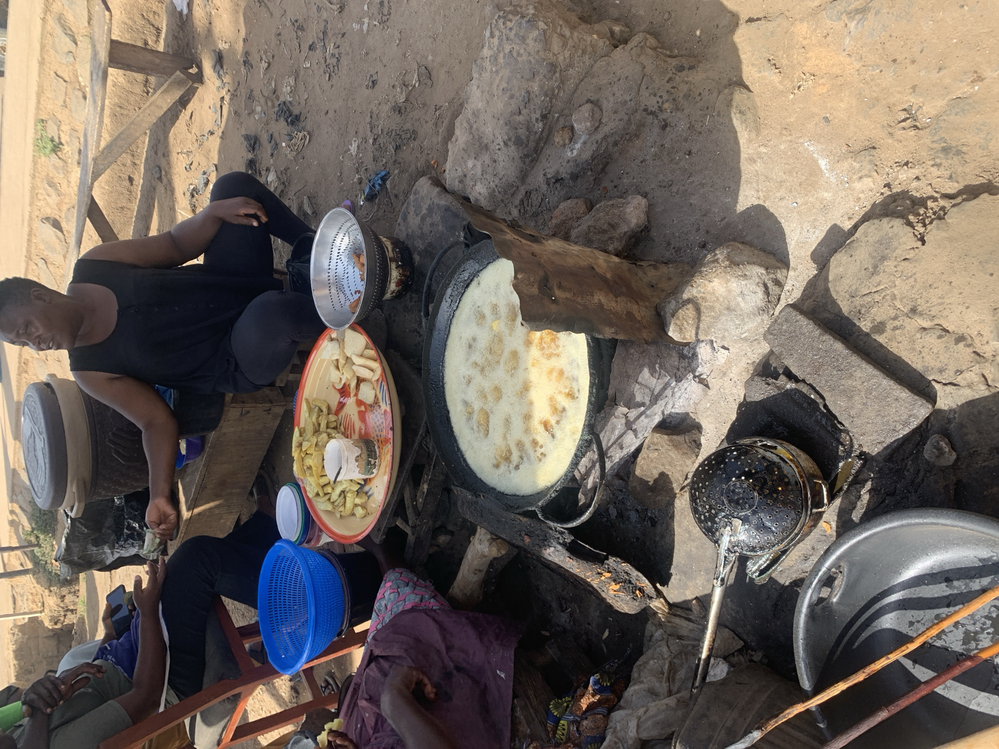 The image depicts an outdoor cooking scene near Rafin Par, Nigeria. A woman wearing a black tank top and leggings sits on a low wooden structure while preparing food. A large wok-like pan sits atop a makeshift stove of rocks and wood, filled with oil in which food is being fried. A plate with raw plantains is next to the wok. Several other people sit nearby, including at least one person using a phone. Cooking tools are scattered around the cooking area. The ground around the area is dusty and rocky.