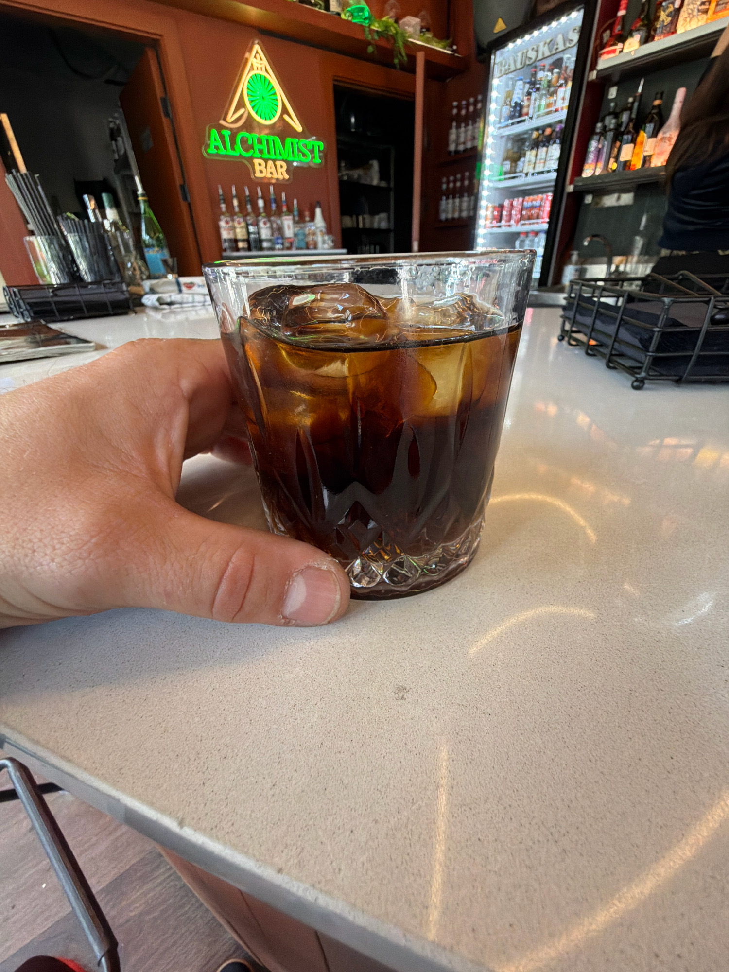 A close-up shot focuses on a hand holding a glass of dark liquid with ice. The setting appears to be a bar, with a neon sign reading "ALCHIMIST BAR" in green and yellow letters visible in the background. Behind the bar, shelves are stocked with various bottles. The overall ambiance suggests a casual and relaxed environment. The image was taken in Riga, Latvia.