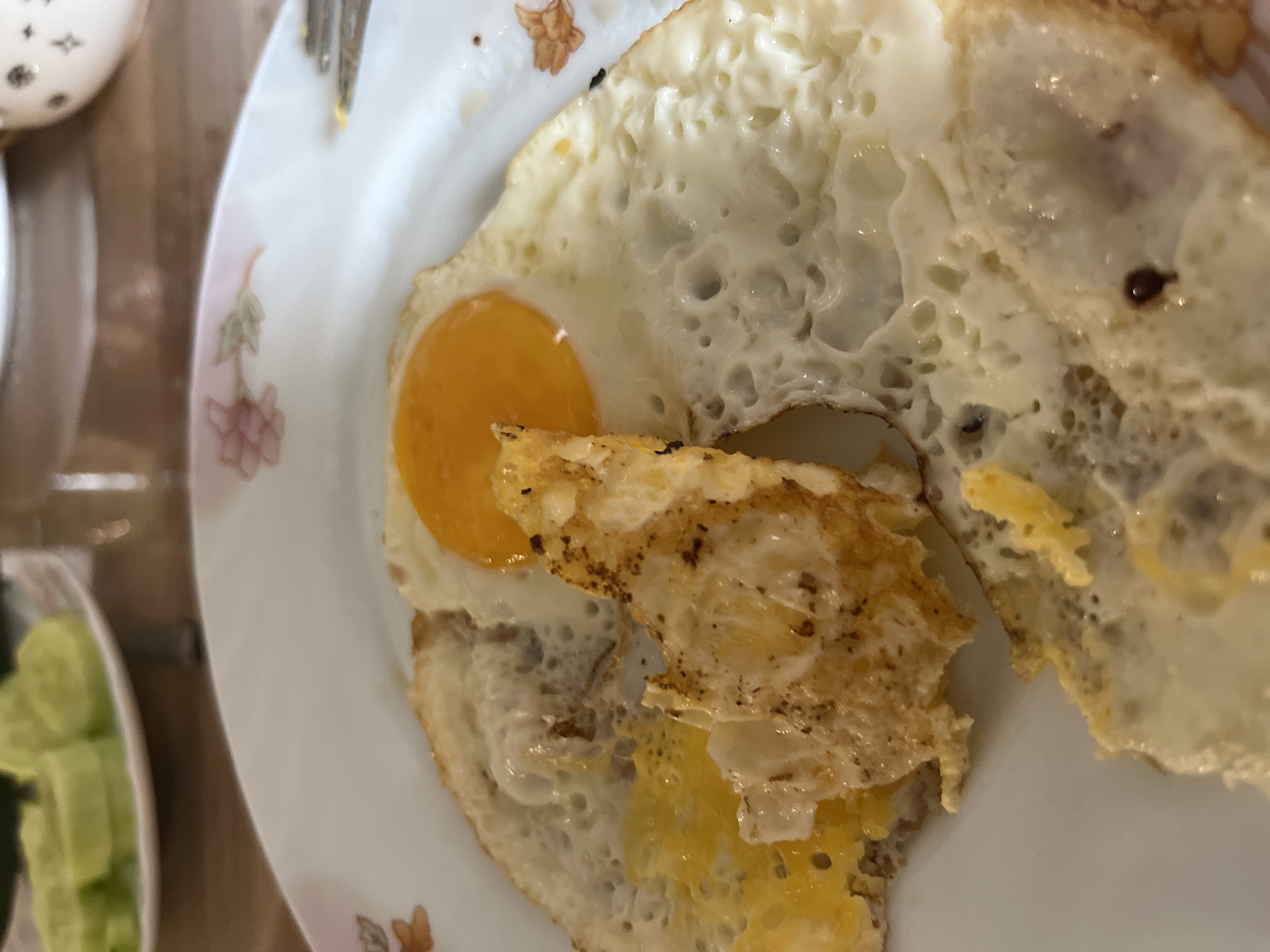 The image shows a close-up of a plate containing fried eggs. One egg has a partially cooked yolk and a bubbled, slightly browned white. A piece of broken egg sits underneath. The plate has a floral pattern. Part of a fork is visible near the edge of the plate. In the background, a dish with sliced cucumbers is partially visible on the left. A second dish is also partially visible. The location, Chaikovsky, Russia, is not discernible from the visual information in the image.