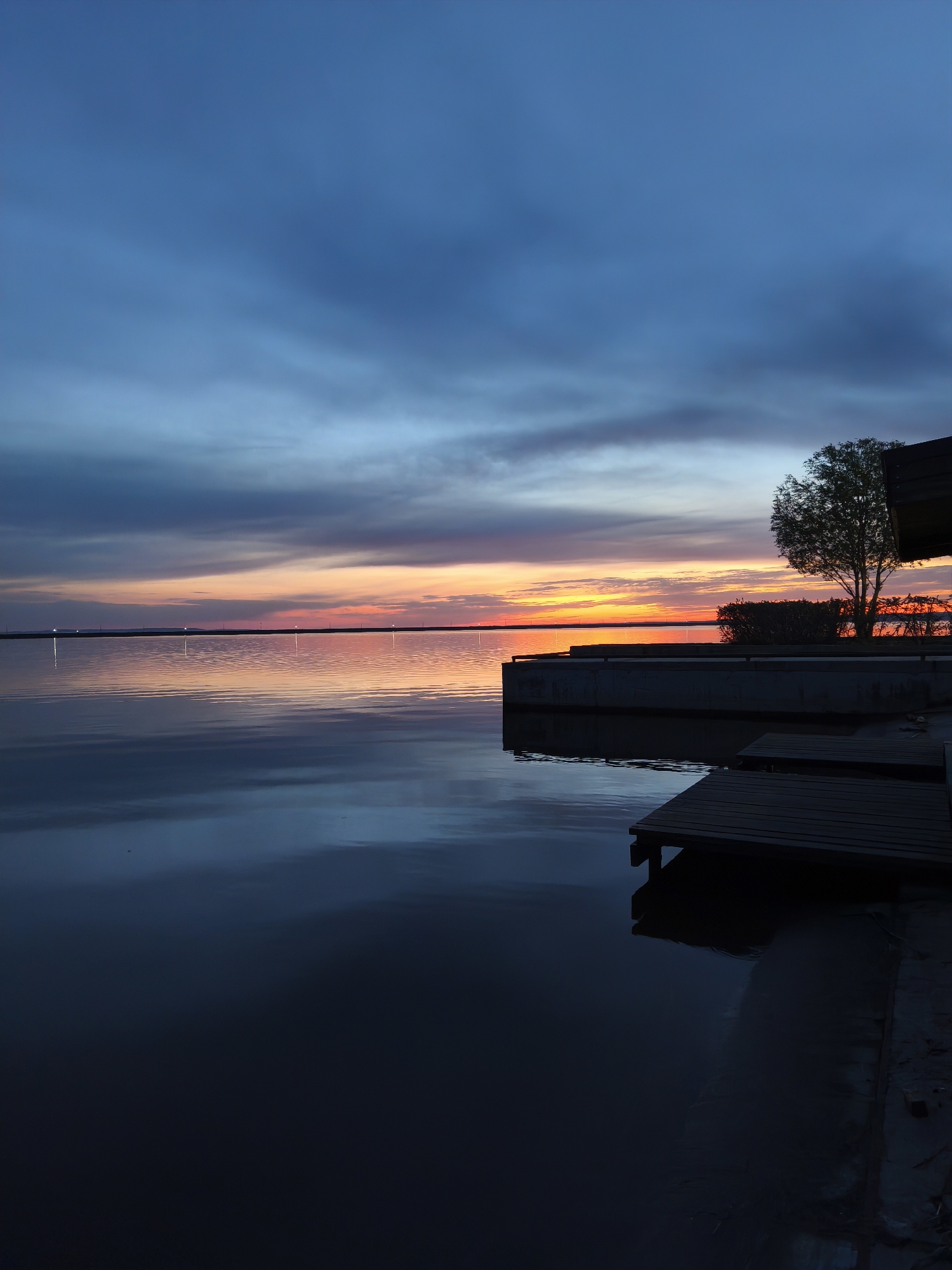 The image depicts a tranquil evening scene at a waterfront location. The sky displays a gradient of colors, transitioning from dark blue at the top to vibrant oranges and reds along the horizon. These colors are reflected in the still water, creating a mirror-like effect.
On the right side of the image, there's a wooden pier extending out into the water. Next to it is a concrete platform with a small tree growing on it, silhouetted against the sunset. The overall atmosphere is serene and peaceful. The scene is located in the city of Chaikovsky, Russia.