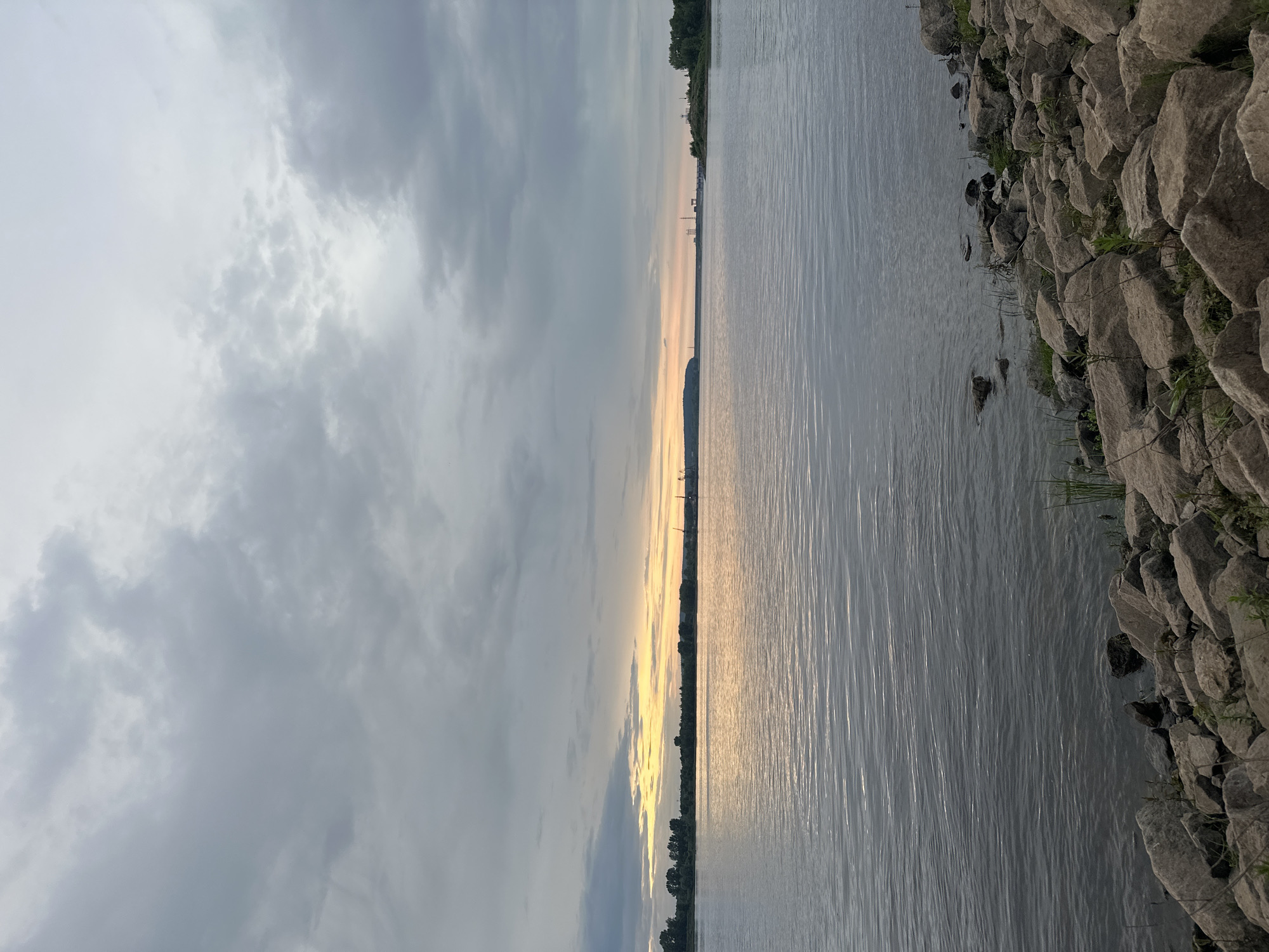 This image depicts a wide, calm river with a rocky shore in the foreground. The sky is overcast with grey clouds, but a band of warm, yellow light is visible on the horizon, suggesting either sunrise or sunset. In the distance, a tree-lined shore is visible, with some industrial structures, including cranes, dotting the landscape. The overall scene is serene and natural, with a hint of human activity on the distant horizon. The location is the Volga River near the city of Noviy, Russia.