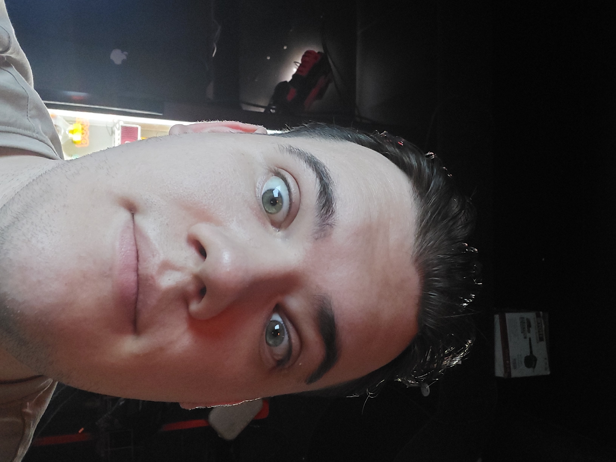 A close-up, slightly rotated self-portrait of a man with fair skin, short dark hair, and light-colored eyes. He has a stubble beard and a neutral-to-slightly amused expression. Behind him, there's a blurred background, suggesting an indoor environment. There appears to be a computer monitor and a cardboard box. There's no indication of the location being Tashkent, Uzbekistan.