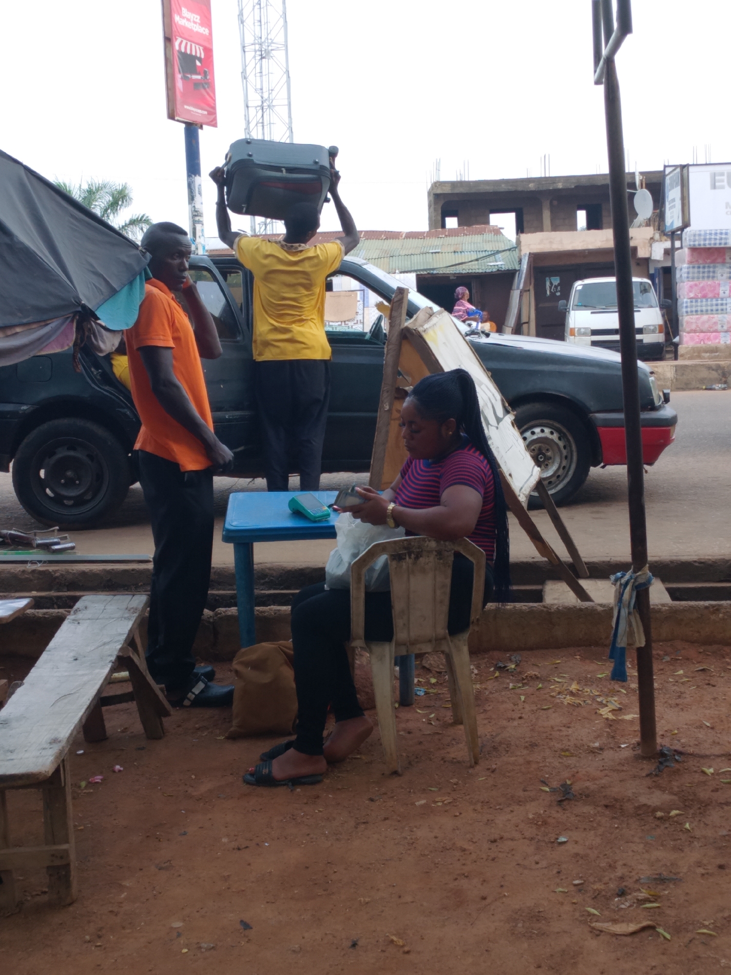An outdoor scene in Sabon Tasha, Nigeria, captures a busy roadside or market area under an overcast sky. The ground is reddish-brown dirt with scattered debris.
In the foreground to the left, a man in an orange short-sleeved shirt and black trousers stands, looking right. Next to him, partially obscured by a large black awning, is a dark-colored car. A man in a yellow t-shirt and dark trousers is seen from behind, skillfully balancing a dark grey suitcase on his head, standing near the car's open door.
To the right, a woman with dark braided hair, tied in a ponytail, sits on a white plastic chair. She wears a red and black striped t-shirt and black trousers, looking down intently at a device in her hands, possibly a mobile phone. Her feet are bare except for dark sandals. On the small blue plastic table in front of her sits a green electronic device, likely a Point of Sale (POS) terminal, suggesting she might be operating a small business or facilitating transactions. A wooden display stand leans next to her, and a rough wooden bench is visible to the left foreground.
In the background, behind the car, various buildings are visible, some appearing unfinished with exposed rebar, and others with corrugated metal roofs. A white van is parked further down the street. A tall metal pole with a red sign is prominent in the upper left. The red sign displays the text "Blayzz Marketplace".
The activity suggests daily commerce and movement, possibly related to travel or roadside vending, capturing a moment of typical street life. No strong emotions are discernible, with individuals appearing focused on their tasks.