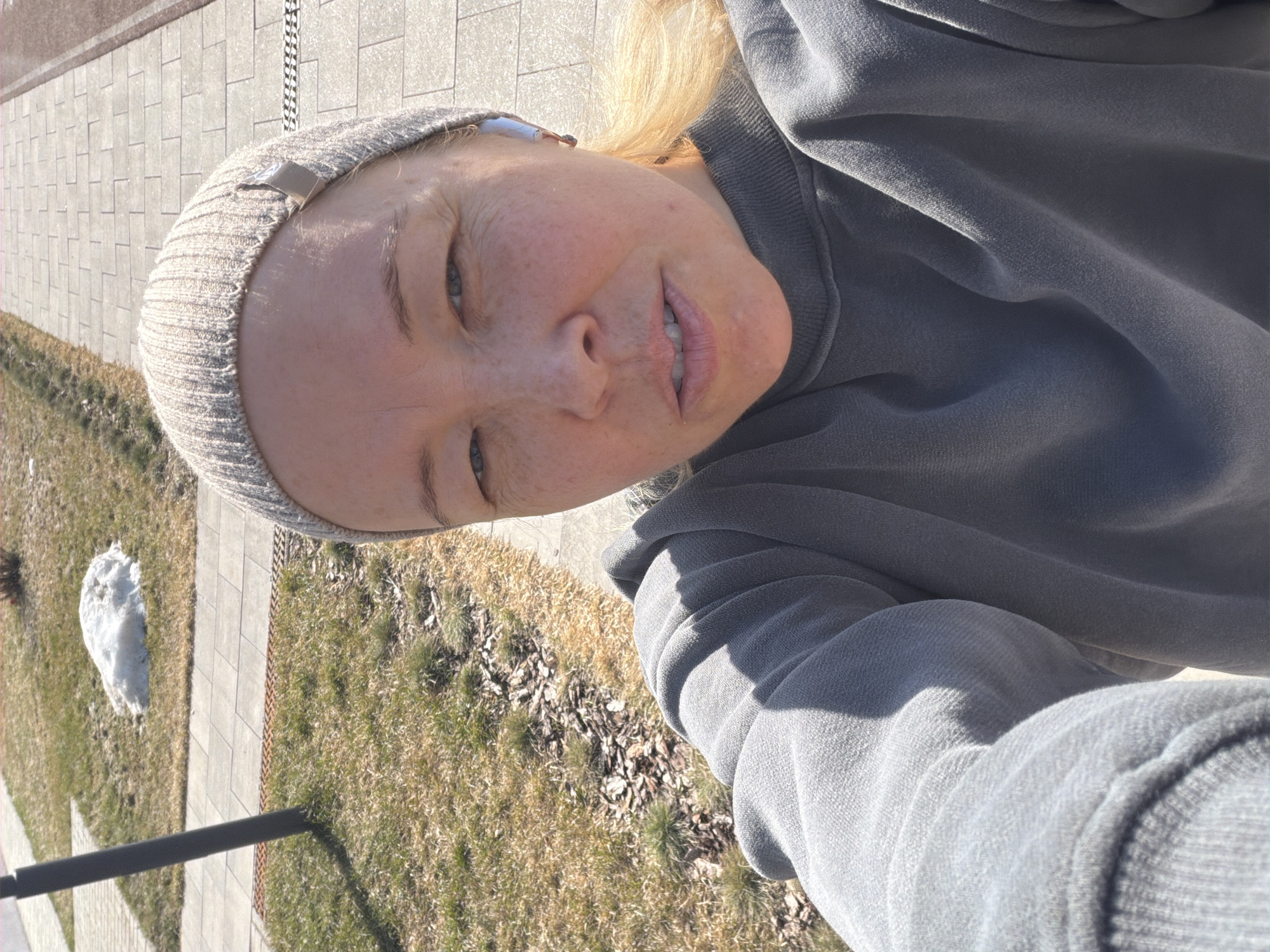 A fair-skinned person wearing a gray beanie and a gray sweatshirt is taking a selfie outdoors. The person's face is tilted towards the camera, and they have blonde hair. The background shows a paved walkway, grass, and some remaining snow. This image appears to be taken in an urban setting, potentially in Pervomaysky, Russia.