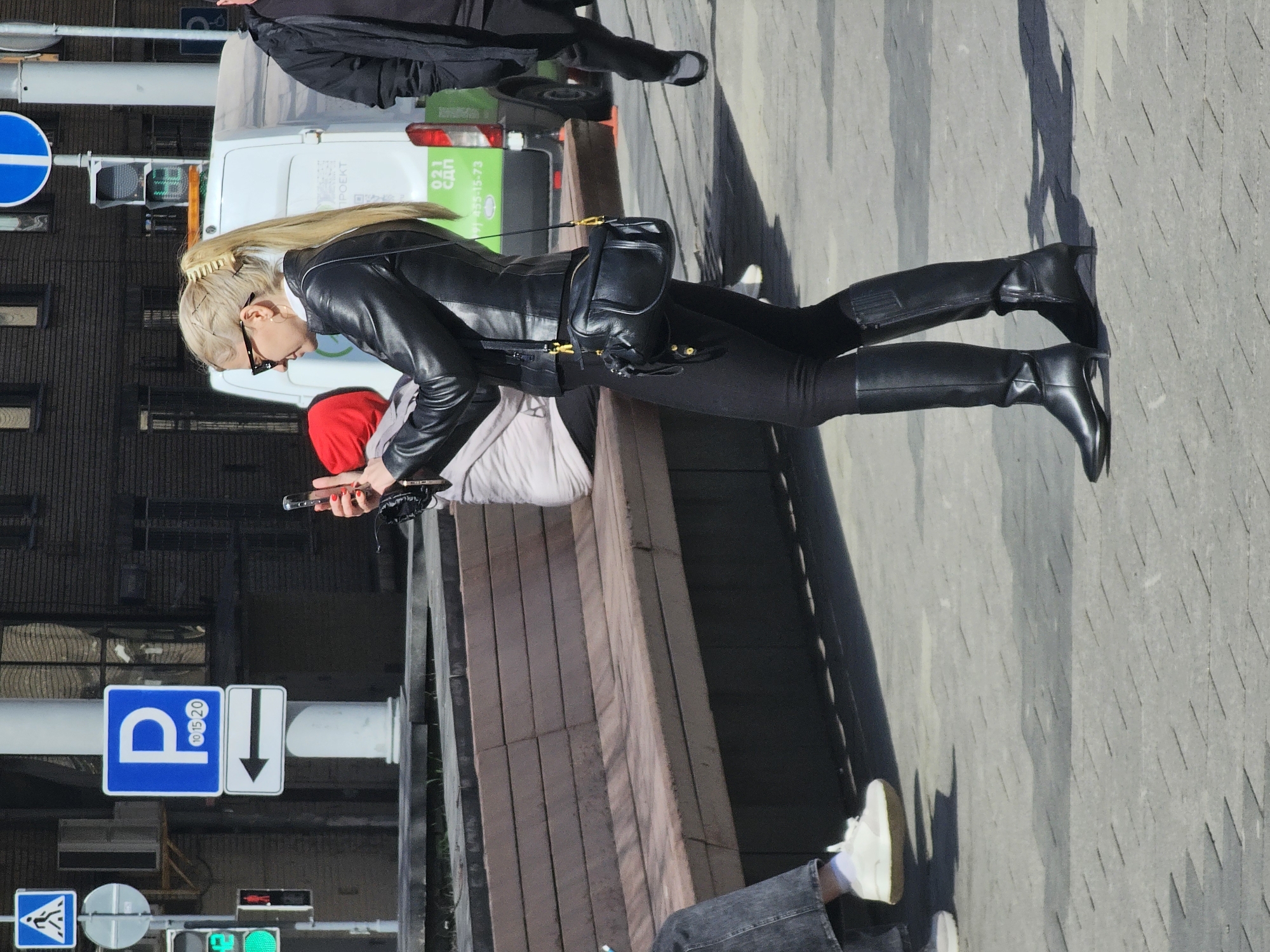 A woman in a black leather jacket, black pants, and black riding boots stands on a city street. She has blonde hair pulled back with a large clip. She wears sunglasses and a small black purse on a strap. She is looking down and holding a cell phone. A second person with a red hat is partially visible. A third person in black clothing is visible in the background. There is a white van with green lettering in the background. A parking sign is visible on a post. A pedestrian crosswalk sign is visible. The street is paved with concrete. Based on the signage and architectural style, the location is in Moscow, Russia.
