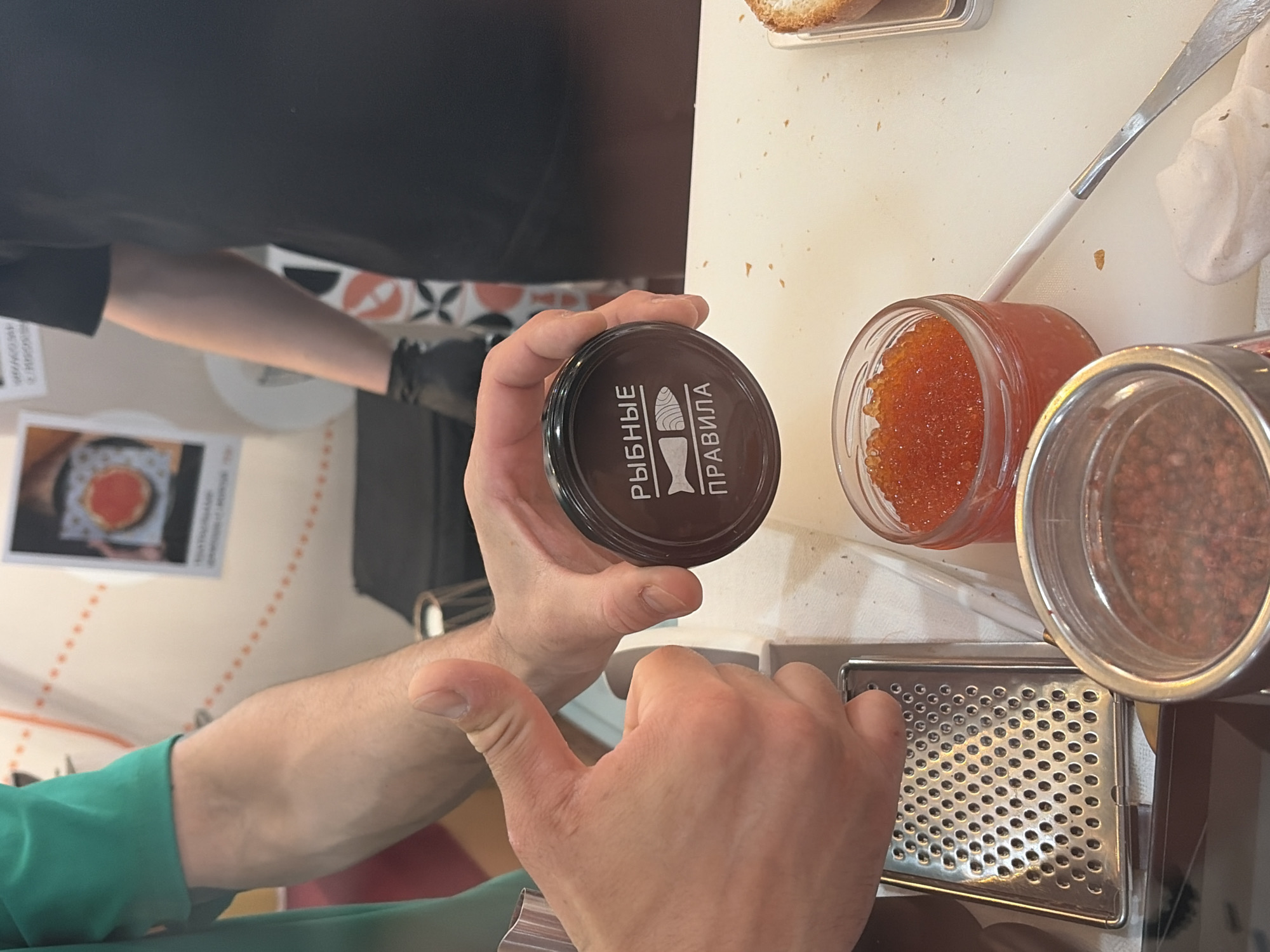 The image shows a close-up of a person's hands holding a small, dark jar with a label that reads "РЫБНЫЕ ПРАВИЛА" and a stylized fish. The person is giving a thumbs-up with their left hand. To the right, there is a small glass jar filled with bright orange caviar and another jar with a reddish substance. A metal grater is visible to the left. In the background, a person wearing a black t-shirt and black gloves can be seen. The scene appears to be indoors, possibly in a kitchen or food preparation area, with some decorative elements visible in the background, suggesting a commercial establishment. The overall context suggests a food tasting or preparation event, possibly related to seafood. The location is not explicitly identifiable from the image itself, but the Cyrillic text on the jar indicates it is likely in Russia or a country with a significant Russian-speaking population.