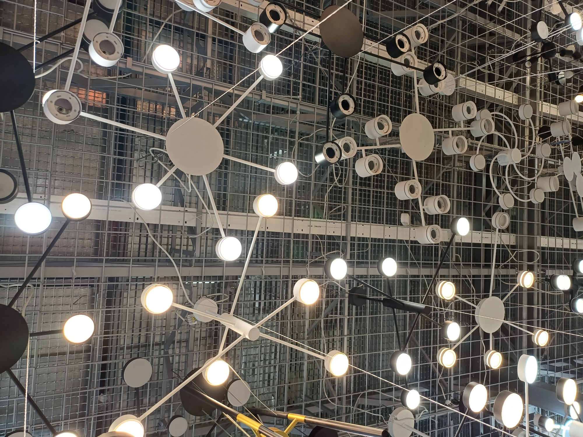 The image shows a collection of modern, geometric chandeliers hanging from a metal grid ceiling. The chandeliers are primarily white and black, with some having spherical light fixtures and others having disk-shaped elements. The overall aesthetic is contemporary and industrial. The lighting fixtures are arranged in various configurations, creating a complex and visually interesting display. The ceiling structure is a typical industrial or commercial type, with metal beams and grid panels. There are no people visible in the image, and no specific location in the city of Boryspil, Ukraine can be determined from the visual information provided.