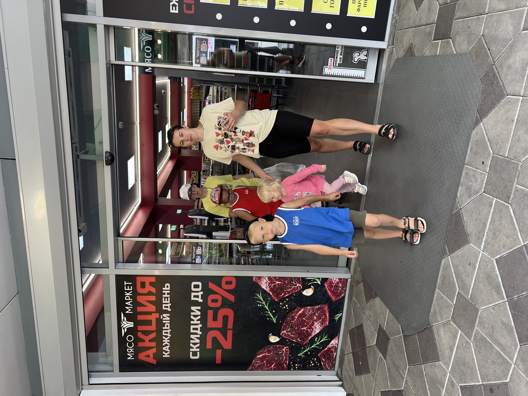 A group of four people, two adults and two children, are walking out of a supermarket. The supermarket has a large sign in Russian advertising meat and discounts. The family is walking towards the camera. The adult woman is wearing a yellow t-shirt with cartoon bears and black shorts. The adult man is wearing glasses and a yellow t-shirt. The boy is wearing a blue tank top and shorts. The young girl is wearing a pink shirt and pink pants. The ground is paved with gray and brown tiles. The supermarket is likely located in Sochi, Russia, based on the Russian text.