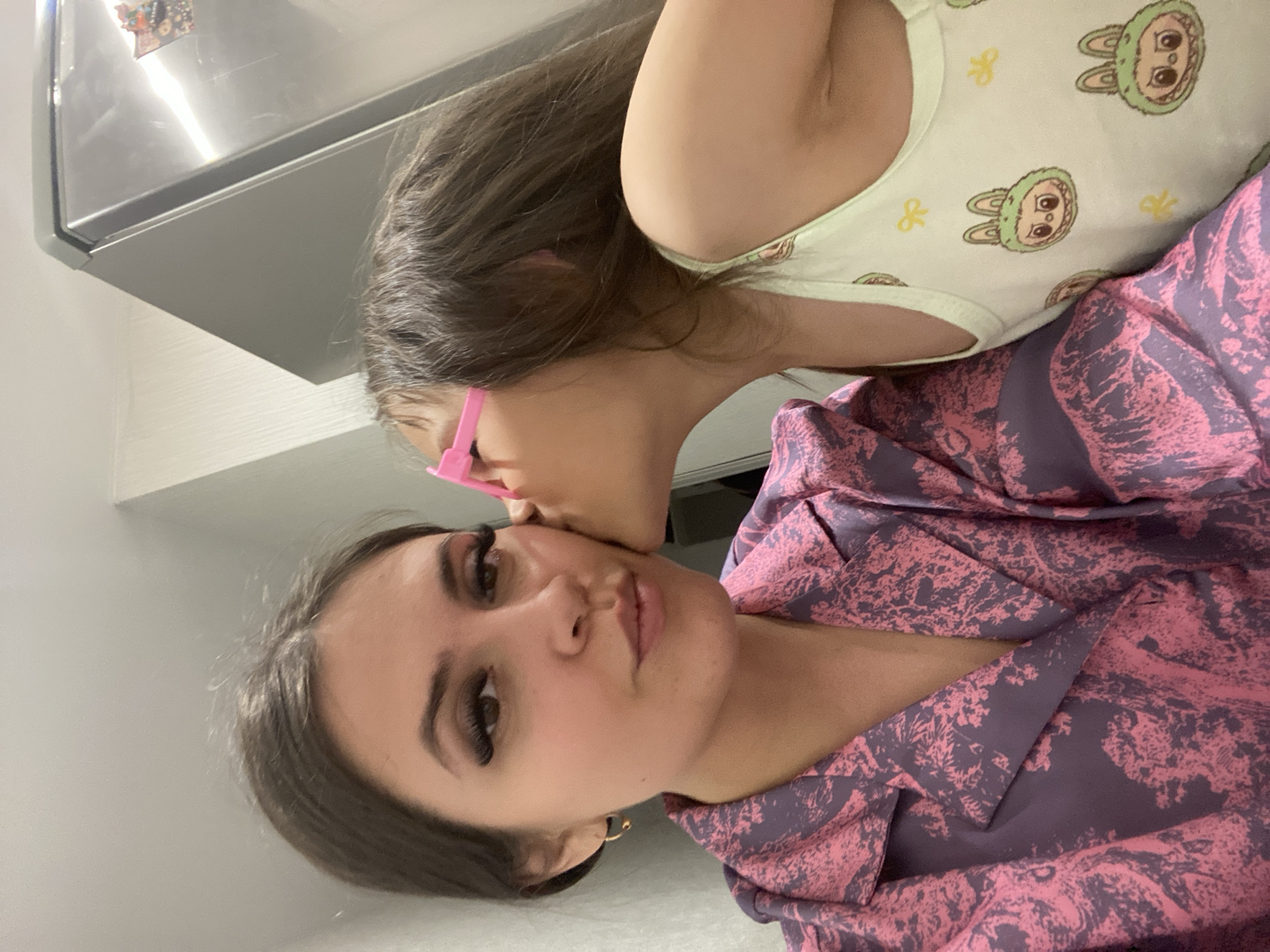 An indoor photograph, rotated 90 degrees counter-clockwise, depicts a mother and child in Chaikovsky, Russia. The child, a young girl, is in the upper right, leaning in to affectionately kiss the woman (presumably her mother) on the cheek. The woman is in the lower left, looking towards the camera with a neutral expression.
The child wears a light-colored tank top or bodysuit patterned with cartoon bunny faces and yellow bows. She has dark hair and appears to be wearing pink-framed glasses or a similar accessory behind her ear.
The woman has dark hair and wears makeup, including prominent false eyelashes. She is adorned with a gold hoop earring in her visible ear and is dressed in a dark purple top or shirt featuring a repeating abstract pattern in pink.
The setting appears to be a home interior. In the background, to the upper left, a shiny, reflective metallic appliance, possibly a refrigerator, is visible with a magnet attached. The rest of the background consists of plain, light-colored walls. The lighting is bright and even, consistent with indoor artificial lighting. No discernible text is visible on any subjects or prominent objects. The scene conveys a warm, intimate moment between the two individuals.