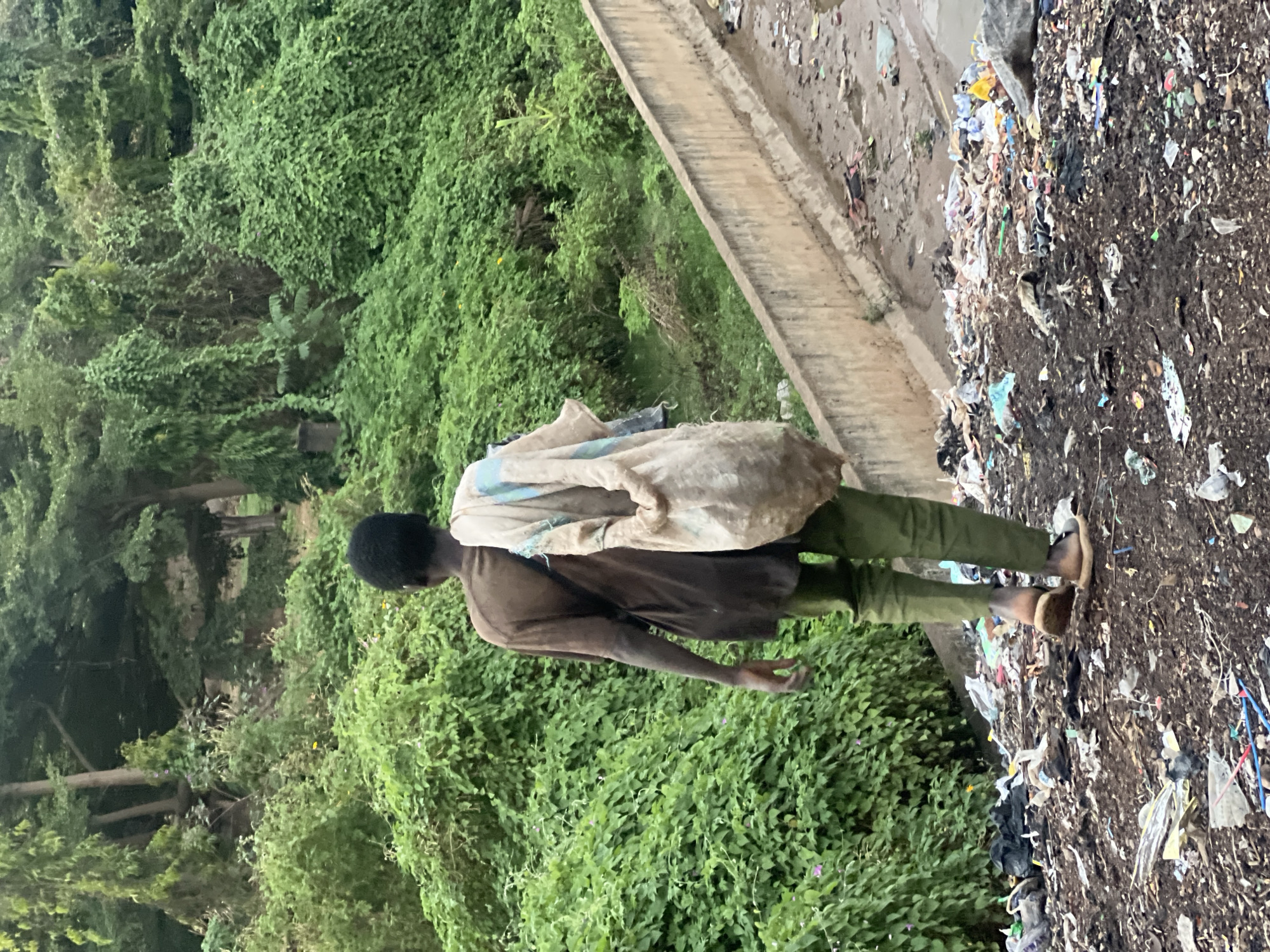 A person stands on a garbage dump in Gura Top, Nigeria. The person is facing away from the camera and has dark hair. They are wearing a brown shirt, green pants, and sandals. They have a large bag over their shoulder. The garbage dump is littered with plastic and other debris. Green vegetation surrounds the garbage dump.