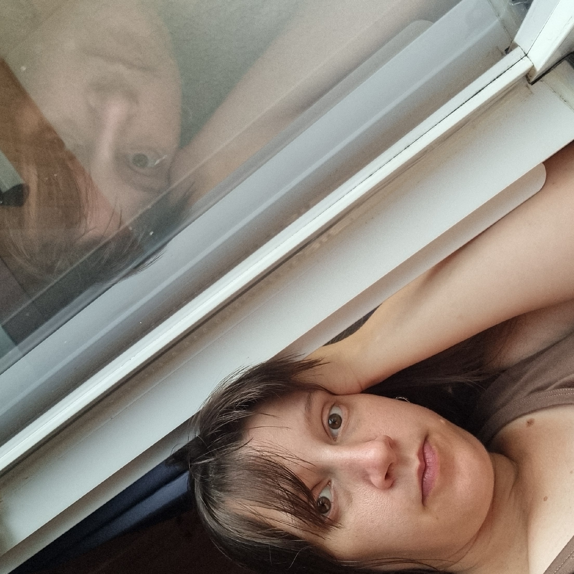 A woman rests her head on her hand, looking directly at the camera. She has brown hair and brown eyes. Her skin is fair. She is wearing a brown tank top. The background is a window frame with some dark fabric to the left. A reflection of her face is visible in the window, slightly blurred and distorted. The scene is indoors, with the window suggesting an apartment or house. There are no specific landmarks visible to pinpoint a location within Dunnae-myeon, South Korea.