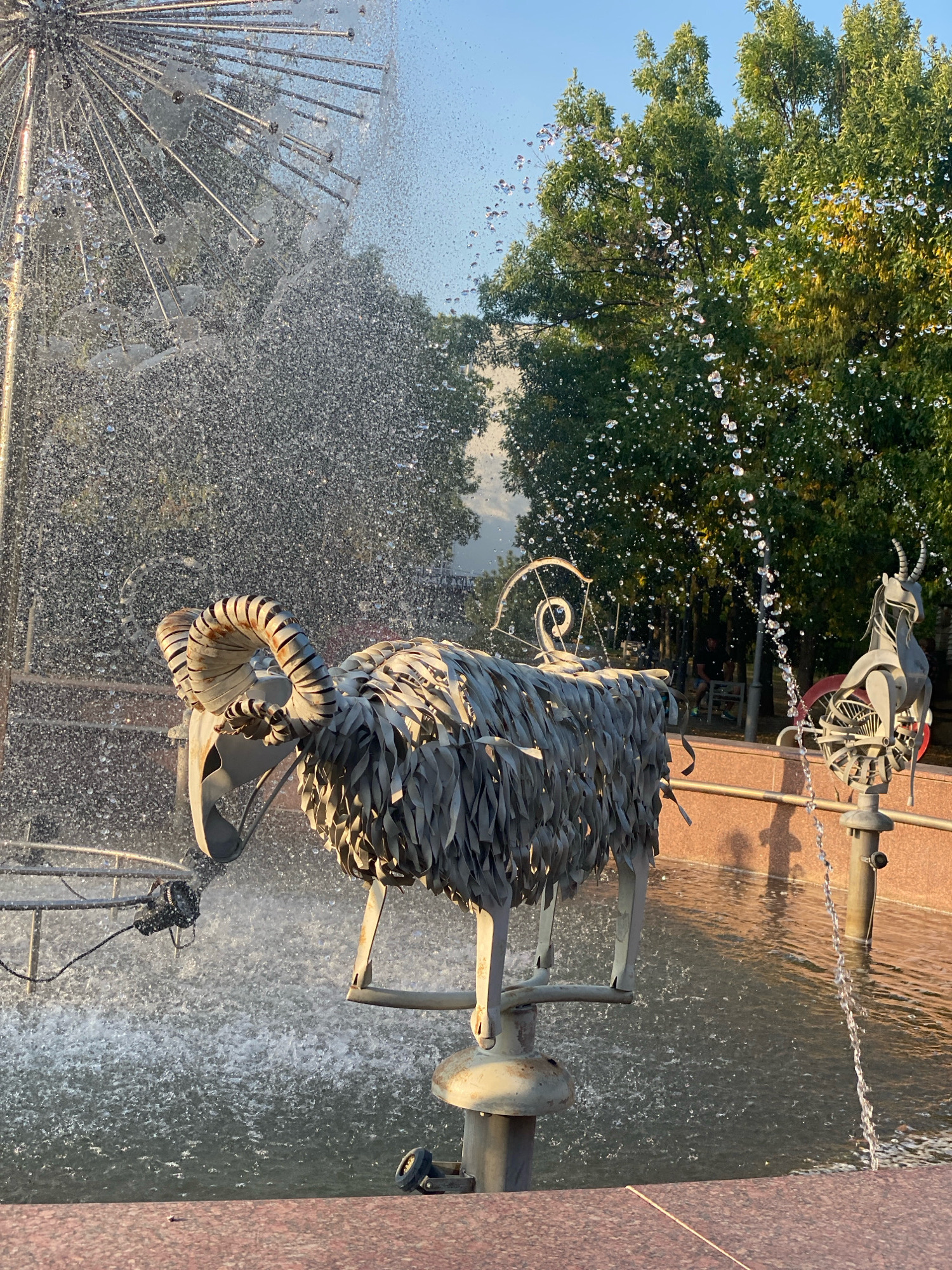 The photograph shows a metal sculpture of a ram with large, curved horns, positioned within a fountain. The ram sculpture is made of overlapping metal strips, giving it a textured appearance. In the background, another metal sculpture, resembling a deer or antelope, is visible. The fountain is active, with water spraying from various points, creating a misty effect. Trees with green foliage are present in the background, suggesting an outdoor setting. The overall lighting indicates it is daytime, with the sun casting shadows.
This sculpture, along with the deer sculpture, is part of the "Water Lily" fountain located in the Rose Valley (Valea Trandafirilor) park in Chișinău, Moldova. The fountain is a prominent feature of the park, and the animal sculptures are a well-known element of its design. The scene captures a moment of the fountain in operation, showcasing the artistic elements and the natural surroundings.