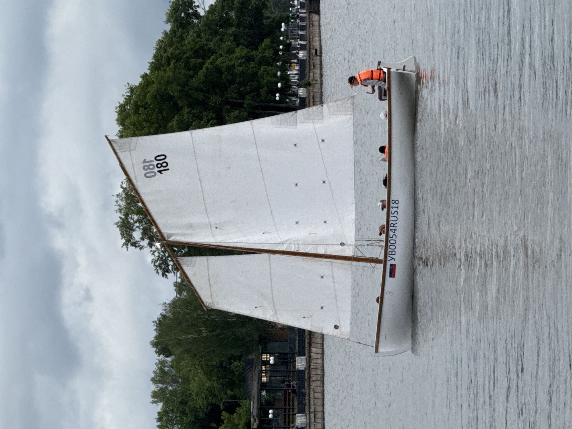 A small, white, gaff-rigged sailboat, registered as YB0054RUS18 and bearing a Russian flag decal, navigates the waters of the Votkinsk Pond in Votkinsk, Udmurtia, Russia, under an overcast sky. The sailboat's main sail displays the numbers "08T" and "180" in black. Approximately four to five individuals occupy the vessel. An adult male, positioned at the stern, wears an orange life vest and a cap while operating the tiller. Three to four additional individuals, some appearing to be children, are seated within the boat's hull. The water is calm with subtle ripples, reflecting the cloudy sky. The background features a tree-lined embankment with a developed shoreline. On the left, a structure resembling a cafe or pavilion with a dark roof and visible interior lighting is partially obscured by trees. Further along the embankment to the right, numerous ornate white streetlights punctuate a paved promenade, which is also lined with dense green trees. The overall scene suggests a leisurely outing on a cloudy day.