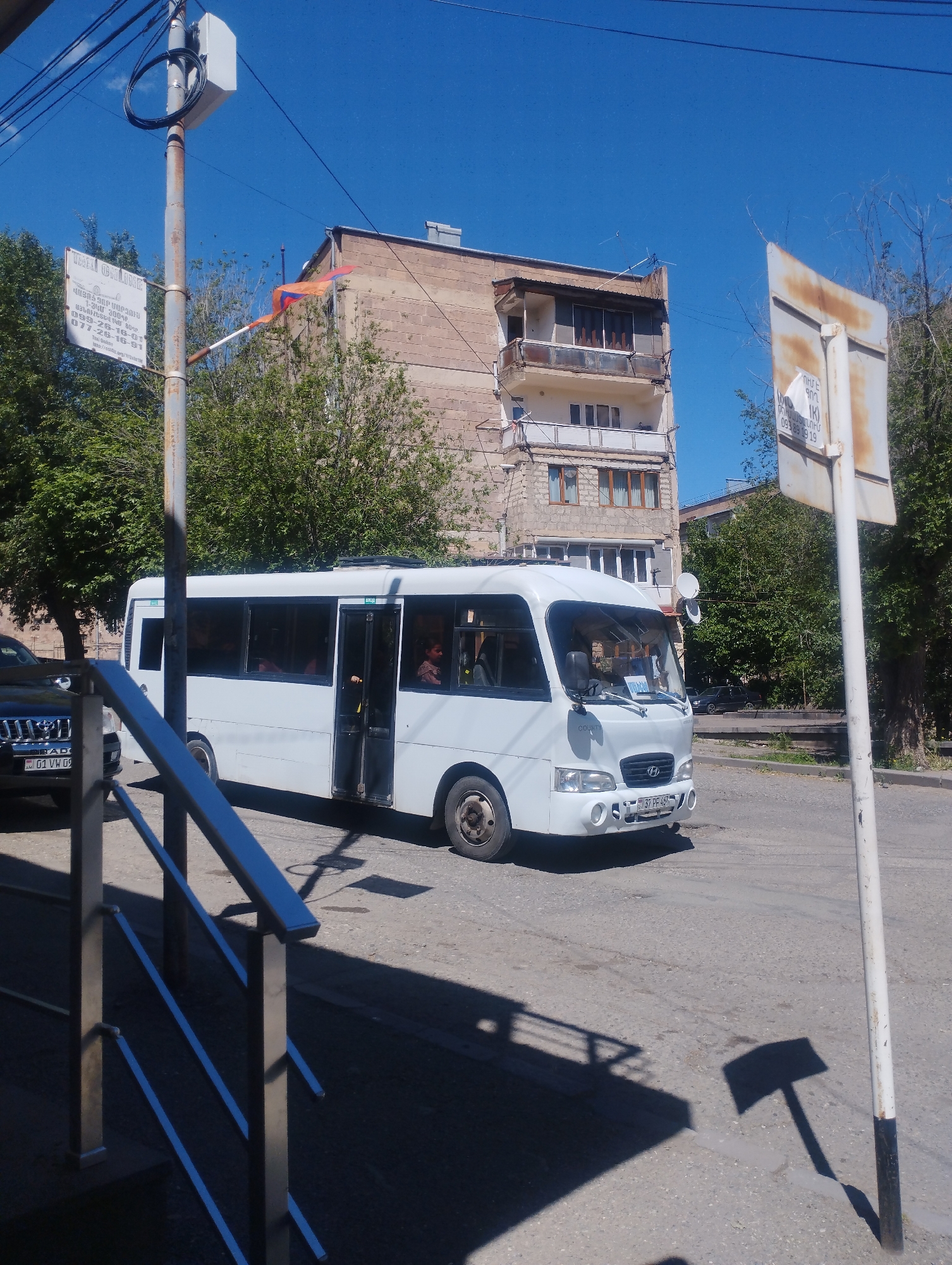 The image shows a white Hyundai County minibus in Yeghegnadzor, Armenia. The bus is on a street next to a sidewalk with metal railings. The bus door is open. A building with balconies is visible in the background. A street sign and a utility pole with a sign are visible on the sides of the bus. There is another car behind the bus, and some people are on the bus. The Armenian flag can be seen flying near the building in the background. The day is sunny.