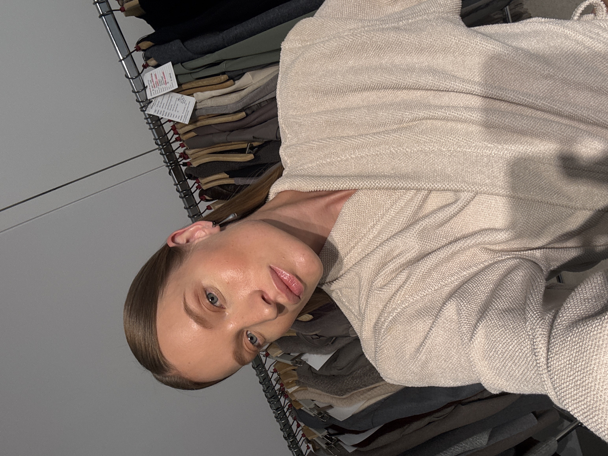 A woman with blonde hair pulled back, wearing a cream-colored robe, is looking towards the camera. She has blue eyes and is wearing subtle makeup. In the background, a rack of clothing hangs, with various garments visible. The scene appears to be indoors, possibly a dressing room or a clothing store. There is no indication of the location being in Oelde, Germany.