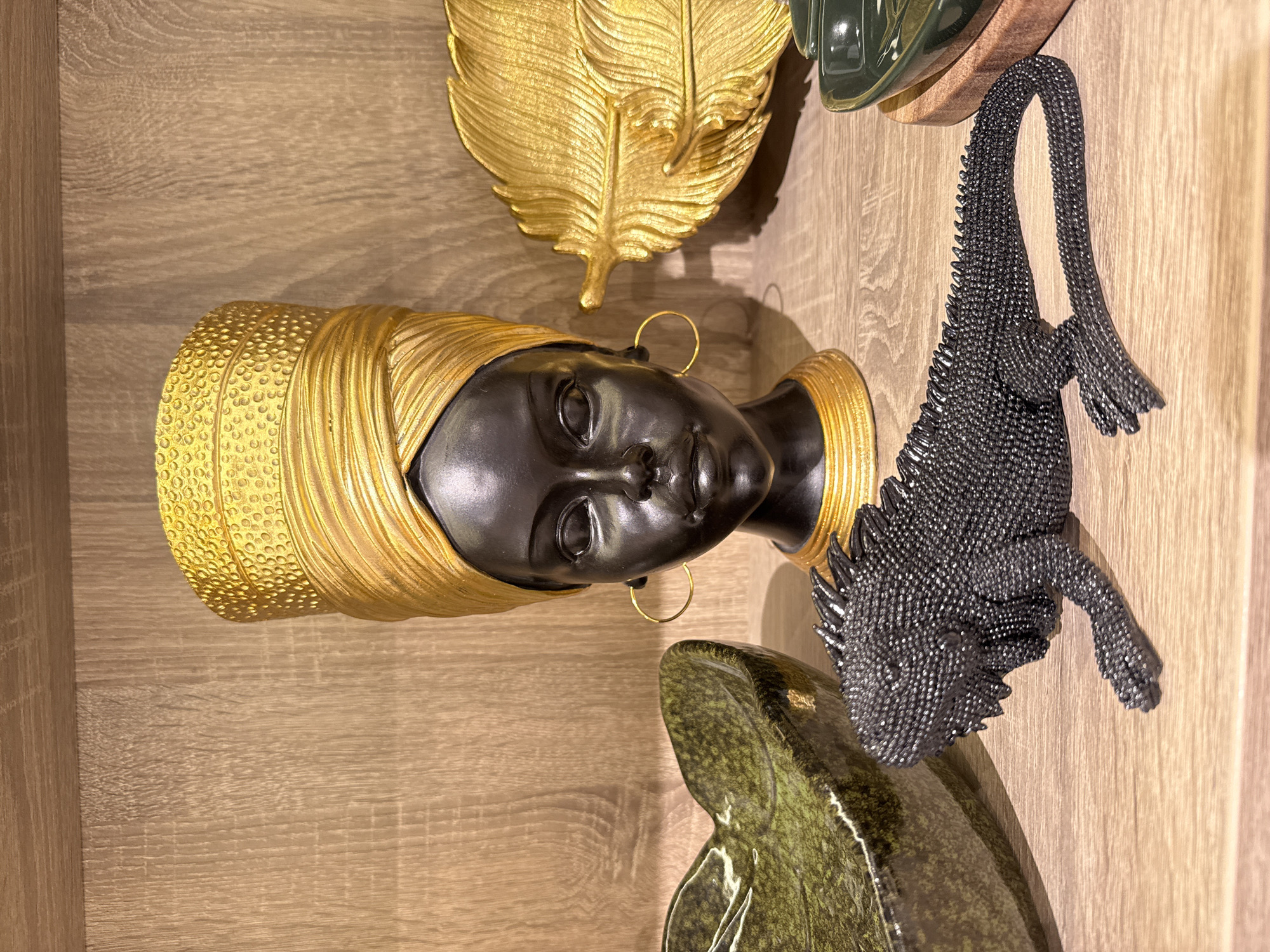 An indoor shot shows a wooden shelf displaying various decorative items. In the center, a bust of a Black woman with a gold turban and necklace is prominently featured. To the left, a textured green bowl is visible, and to the right, a large golden feather decorative piece. In the foreground, a black iguana figurine with a textured surface rests on the shelf. The scene is well-lit, highlighting the textures and colors of the objects. The location in Izhevsk, Russia, cannot be determined from the image itself.