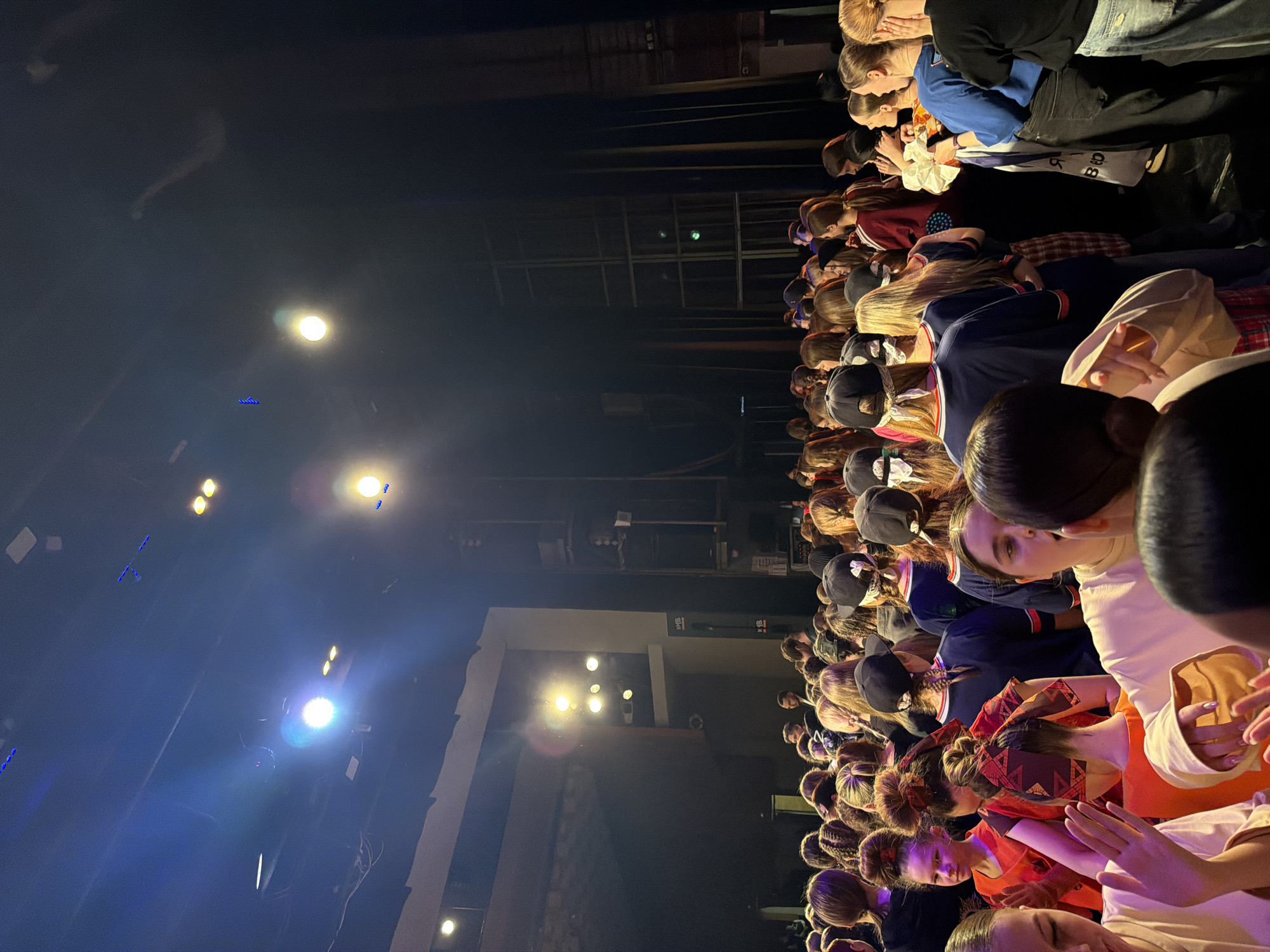 A vertically oriented, wide shot captures a large gathering of people, primarily girls and women, inside a dimly lit auditorium or theater. The viewpoint is slightly elevated, looking down and across the rows of individuals. The subjects are seated closely together, predominantly facing forward. Many are wearing hats or ribbons. In the upper right of the frame, some people are dressed in more casual attire, seated in rows behind the main group. In the upper right of the frame, some people are dressed in more casual attire, seated in rows behind the main group.
The environment is stark, with spotlights and stage lighting illuminating portions of the scene. The backdrop consists of dark curtains and architectural elements, indicating an indoor venue. The location is Perm, Russia. The interaction between the subjects is subtle; they appear attentive, possibly watching a performance or presentation on stage. The actions observed are mainly of sitting, looking, and perhaps quietly communicating.