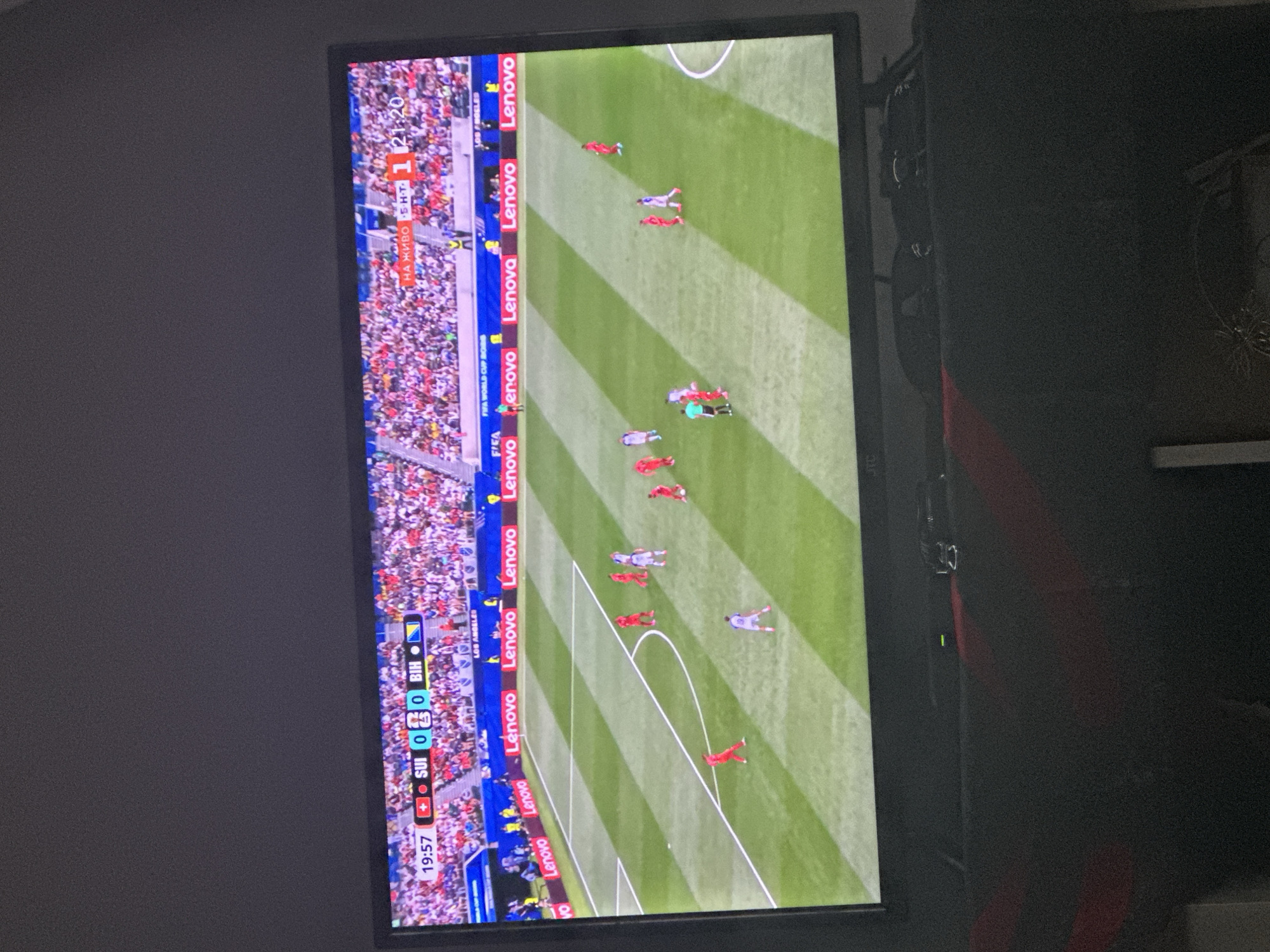The image shows a television screen displaying a live soccer match. The score is 0-0 between Switzerland (SUI) and Bosnia and Herzegovina (BIH) at 19:57. The game is being played in a stadium with a crowd in the background. There are advertisements for "Lenovo" and "FIFA WORLD CUP RUSSIA" visible on the screen. The television is placed on a dark surface, possibly furniture, in a dimly lit room. The overall scene suggests a casual viewing of a sporting event. There is no indication of the specific location in the city of Mainburg, Germany.
