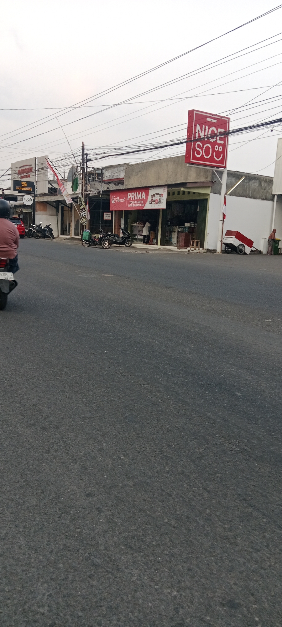 A street view captures a row of shops in Maguwoharjo, Indonesia. A red sign with "NICE SO" and a smiley face is prominently displayed above one shop. Various motorcycles are parked in front of the shops, and a person is seen exiting one of them. The shops appear to be a mix of businesses, including one labeled "Vapehouse," another with "Parrot PRIMA TOKO PLASTIK DAN BAHAN KUE" (plastic and cake ingredients store), and others with less clear signage. A small red and white cart is parked near one of the buildings, and a person is standing next to a trash bin. The road in the foreground is asphalt, and electrical wires crisscross overhead. The sky is overcast.