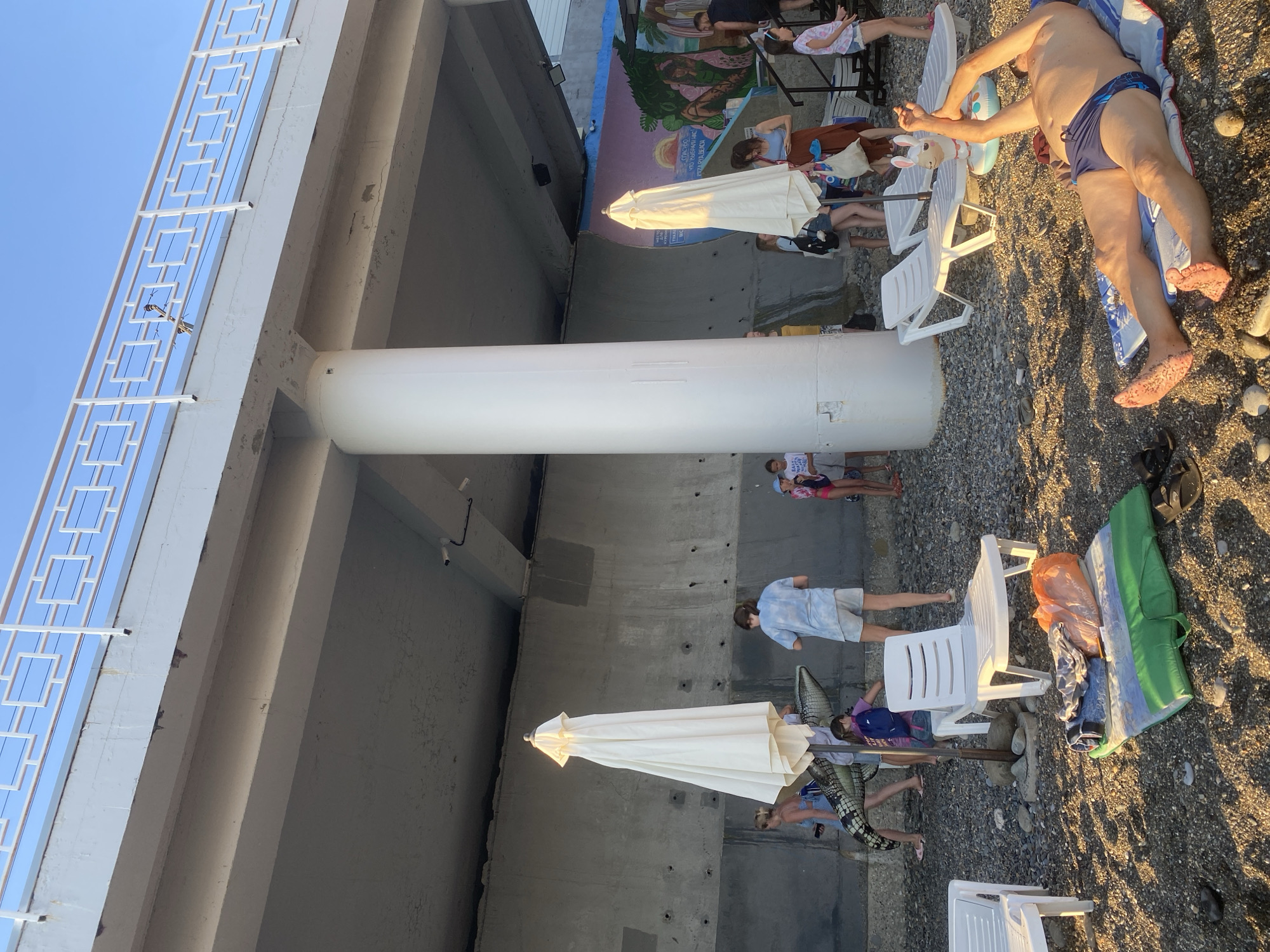 This media file captures a scene on a pebble beach, likely in Sochi, Russia, during what appears to be the late afternoon or early evening, given the warm, low-angled sunlight. The setting is under a large concrete structure, possibly a bridge or overpass, which casts shadows and provides a backdrop.
Several individuals are present, mostly lying down or sitting on beach chairs, enjoying a relaxed day at the beach. In the foreground, a man in blue swimming trunks is lying on a towel, his arms outstretched. Further back, people are scattered amongst white plastic beach chairs and closed, off-white beach umbrellas. Some individuals appear to be sunbathing, while others seem to be resting or socializing. One person is wearing a light blue shirt and shorts, and another is in a patterned top and shorts. A closed white beach umbrella is prominently positioned.
Behind the beachgoers, a colorful mural is visible on the concrete wall of the overpass, depicting a tropical landscape with greenery and a sun. The sky above is a clear, bright blue. The ground is covered in grey pebbles, characteristic of many Black Sea beaches. There is a sense of leisure and relaxation pervading the scene. The overall atmosphere is peaceful, with people enjoying the coastal environment under the warm sunlight.