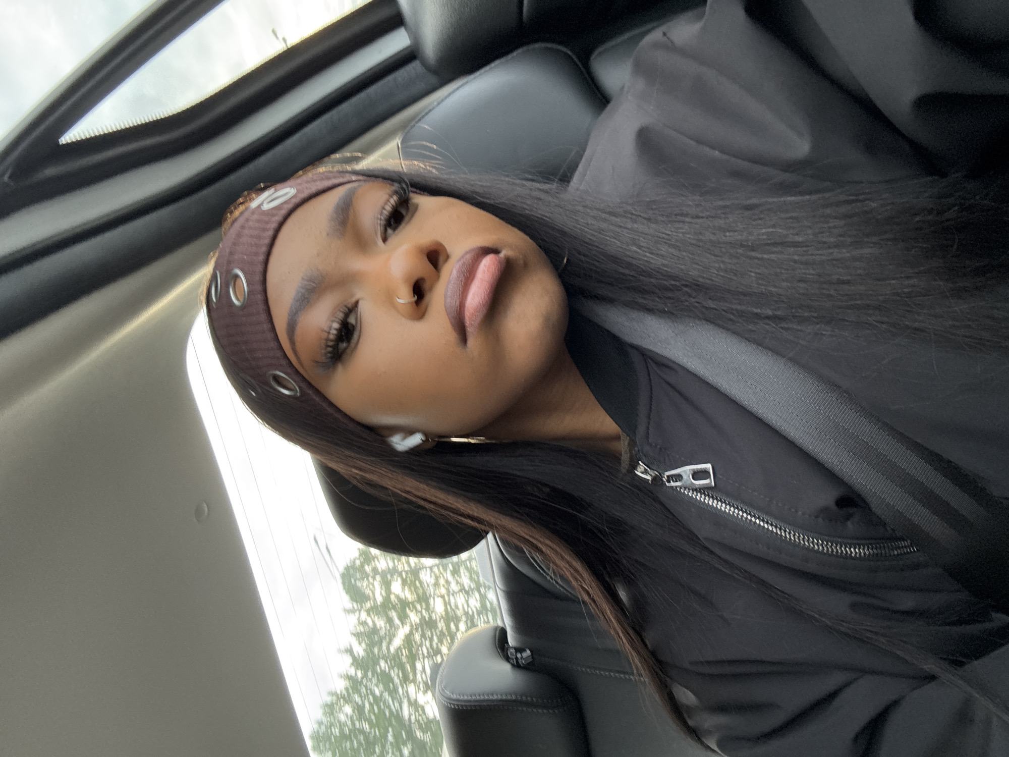 A medium-angle, eye-level shot shows a young, dark-skinned woman seated in a car, wearing a brown headband with silver grommets. The woman is wearing a black bomber jacket and a seatbelt. The background shows part of the car's interior, including the seat, headrest, and sunroof, along with blurred trees and sky visible through the windows. The image appears to be taken in a car in any city, and there's no evidence in the picture that suggests the city is Moscow, Russia.