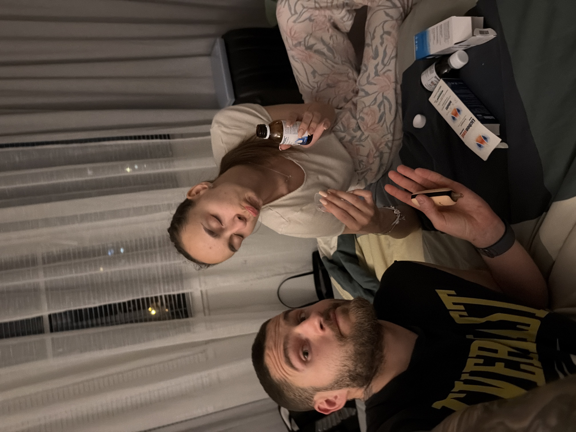 The image captures two individuals, a woman and a man, in an indoor setting, likely a bedroom or living room. The perspective is from above, with the image rotated such that the woman is positioned at the top and the man at the bottom.
The woman, with dark hair, is reclining, wearing a light-colored top and floral-patterned pajama bottoms. She is actively engaged in preparing or taking medicine, holding a small brown bottle with a white label in her right hand and a small, clear measuring cup or spoon in her left hand. Part of the brand name "KCEKC" is visible on the bottle. Her nails are manicured.
The man, sporting a beard, is also reclining, wearing a black t-shirt with the word "EVERLAST" emblazoned in yellow capital letters across the chest. He looks upwards, seemingly observing the woman or the general situation. In his left hand, he holds a small, light-colored object that resembles a vape pen or a similar personal device. A dark-colored watch is visible on his left wrist.
Between and around the individuals, particularly near the man, several pharmaceutical products are visible: a white and blue box, a white bottle, and a white tube or box with the brand name "Lichtensud" clearly readable.
The background shows light-colored curtains and what appears to be horizontal blinds or an exterior structure, through which faint artificial lights are visible, indicating the time of day is evening or night. A ventilation grate or air conditioning unit is also present near the window area. The overall scene conveys a moment of rest or recuperation, possibly due to illness, in Sofia, Bulgaria.