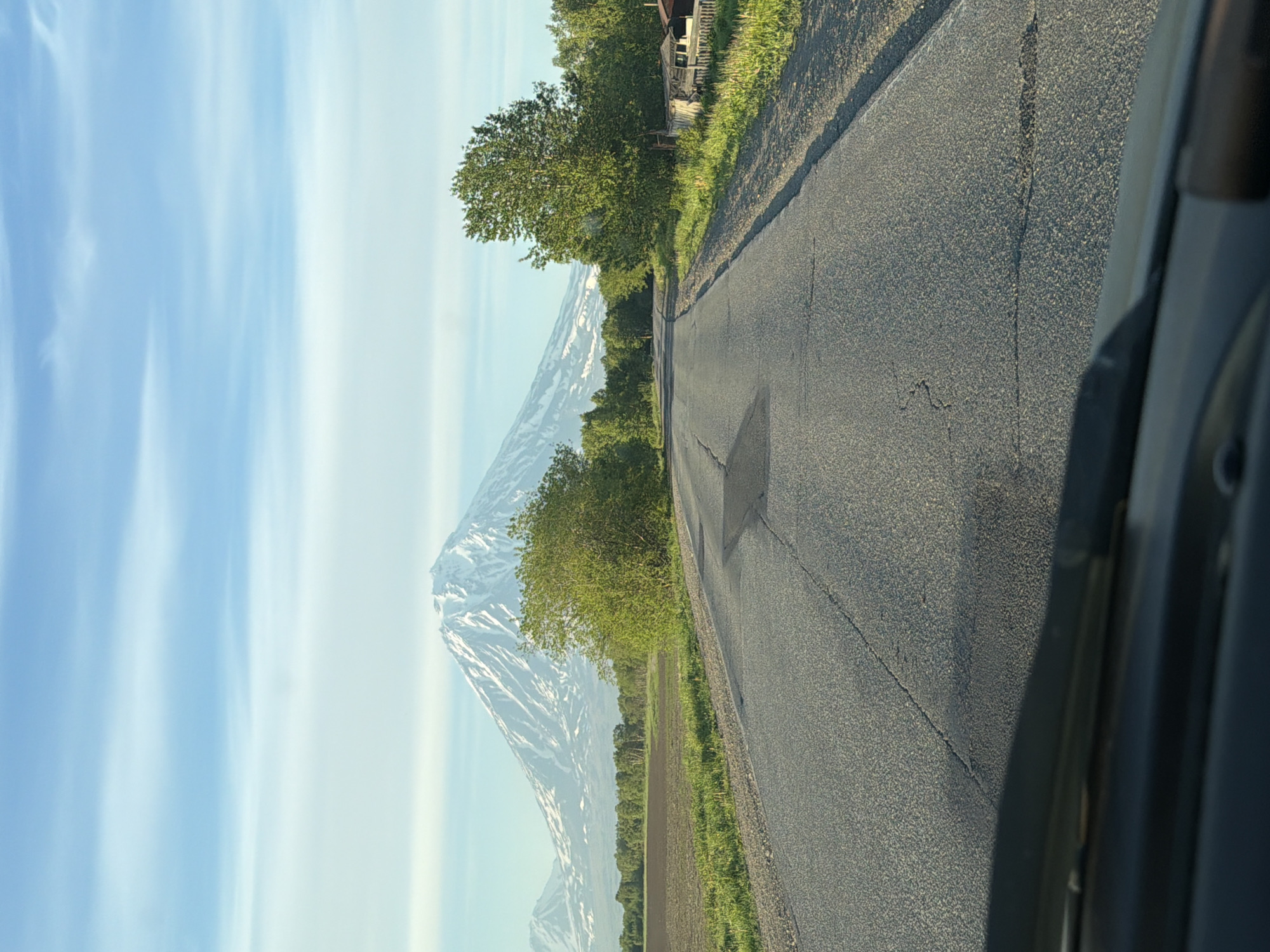 The media file captures a view from a vehicle, looking out at a scenic landscape. The scene is dominated by a snow-capped mountain, likely a volcano, with its slopes displaying streaks of white snow against darker rock or soil. The mountain is framed by lush green trees and vegetation, suggesting a temperate climate. To the right, an asphalt road curves away from the viewer, its surface showing signs of wear and tear, including visible cracks. The roadside is lined with grass and more trees, and in the distance, a wooden structure, possibly a house or shed, with a fence, is partially visible. The sky is a vibrant blue, streaked with wispy white clouds that create a sense of depth. The overall lighting suggests it is daytime, possibly morning or late afternoon due to the soft quality of the light. The image is oriented vertically, implying it was taken with the camera held in portrait mode. The view gives a sense of travel and exploration, with the road leading towards the majestic mountain. No people are visible in the frame. The setting appears to be rural or semi-rural, given the presence of the natural landscape and the solitary structure. The location context provided is Yelizovo, Russia, which aligns with the possibility of volcanic mountains being present in the region.