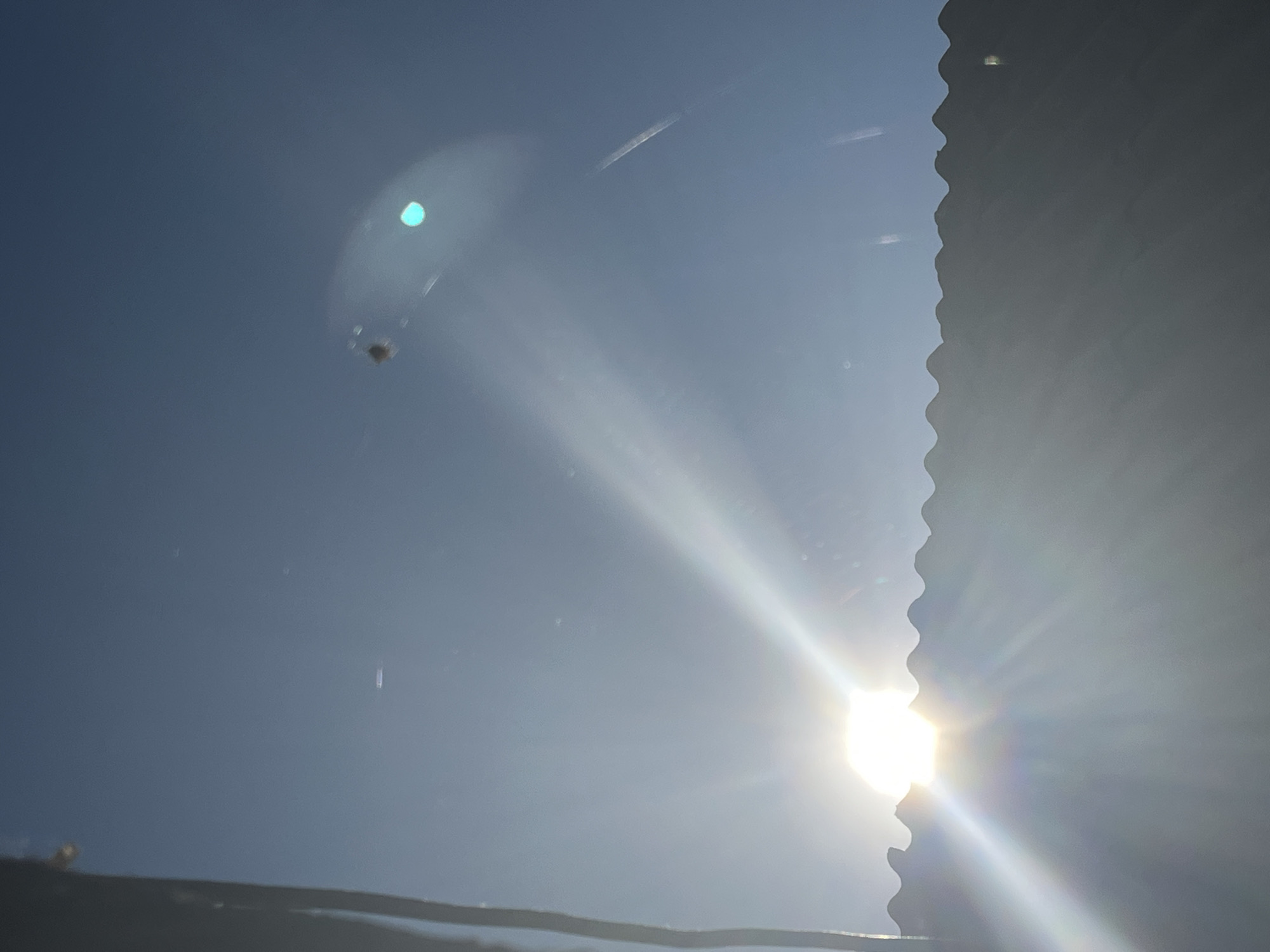 The image shows a bright sun with rays shining through a corrugated surface, likely a rooftop or fence, casting a glare. The sky is clear and blue. In the upper part of the image, a small, dark object, possibly a spider, is visible, suspended by a thin thread against the sky. There are also lens flares and reflections on what appears to be a window or glass surface, adding a layer of abstraction to the scene. The overall impression is one of a bright, sunny day with an unexpected detail in the form of the suspended spider. The image is taken from a low angle, looking up towards the sky. The location in Krasnyye Barrikady, Russia, is not visually evident from the image itself.