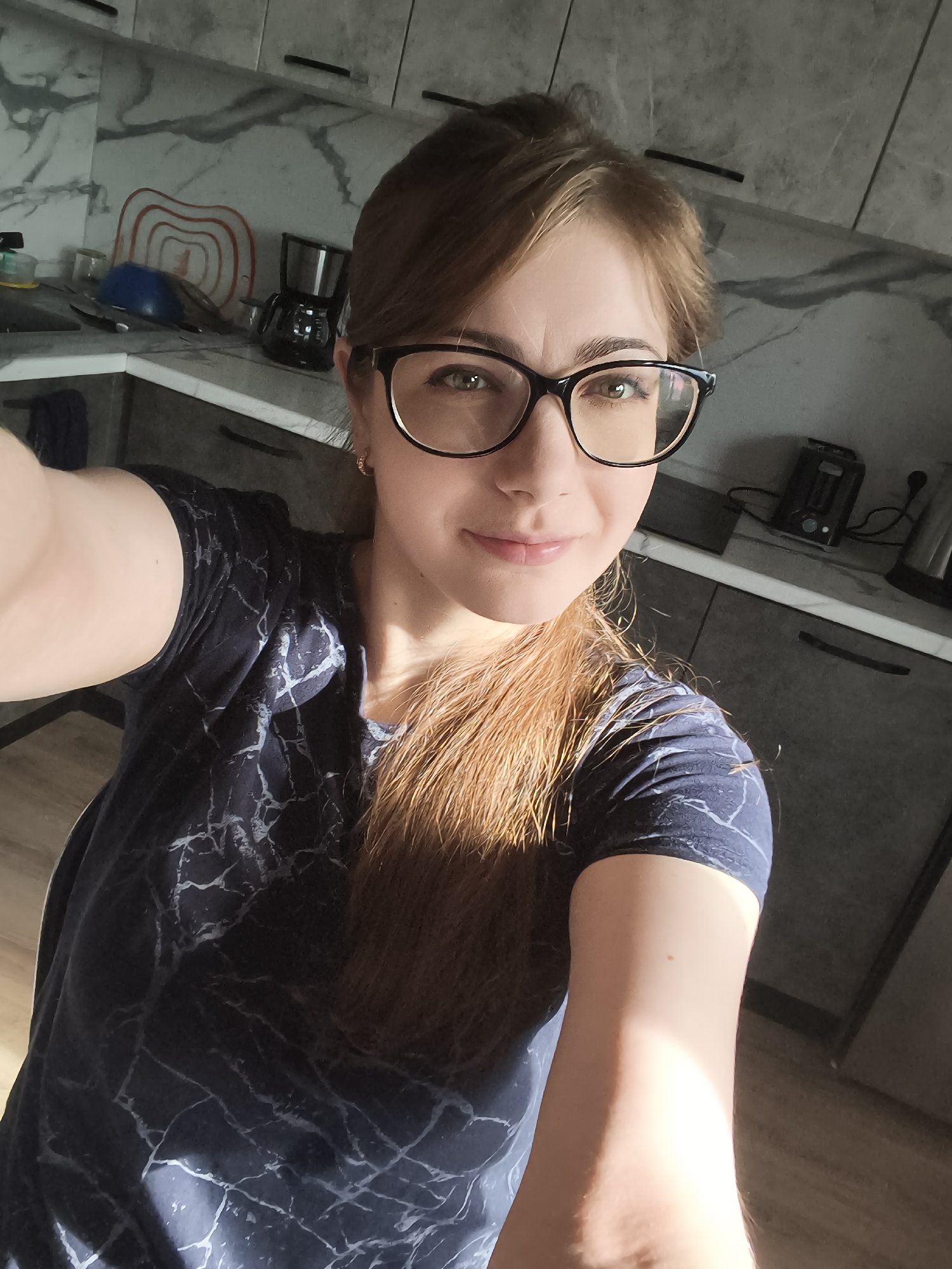 A woman with auburn hair and glasses is taking a selfie in a kitchen. The kitchen features gray cabinets, a marble-patterned backsplash, and a white countertop. Visible appliances include a coffee maker and a toaster. The woman is wearing a dark blue t-shirt with a marble-like pattern. The image was taken in the city of Podkletnoe, Russia.