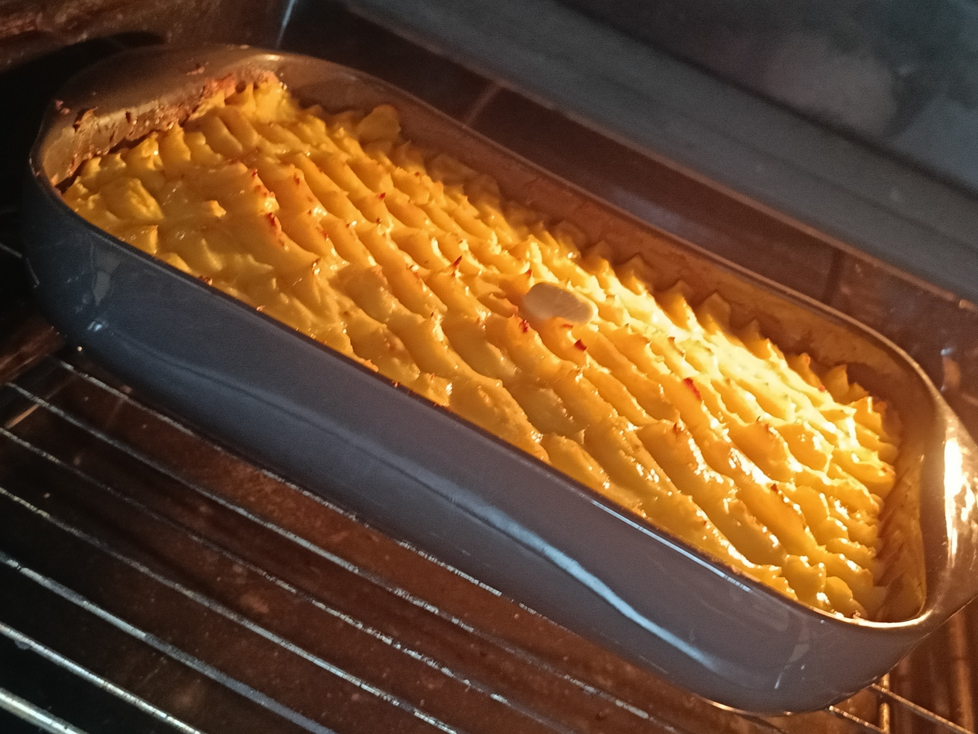 The image shows a shepherd's pie baking in an oven. The shepherd's pie is in a grey oblong baking dish. The topping appears to be piped mashed potatoes. The dish is on a rack inside a lit oven. There are no people visible in the image. The location is not evident from the image. There is no information to indicate this image was taken in Chaikovsky, Russia.