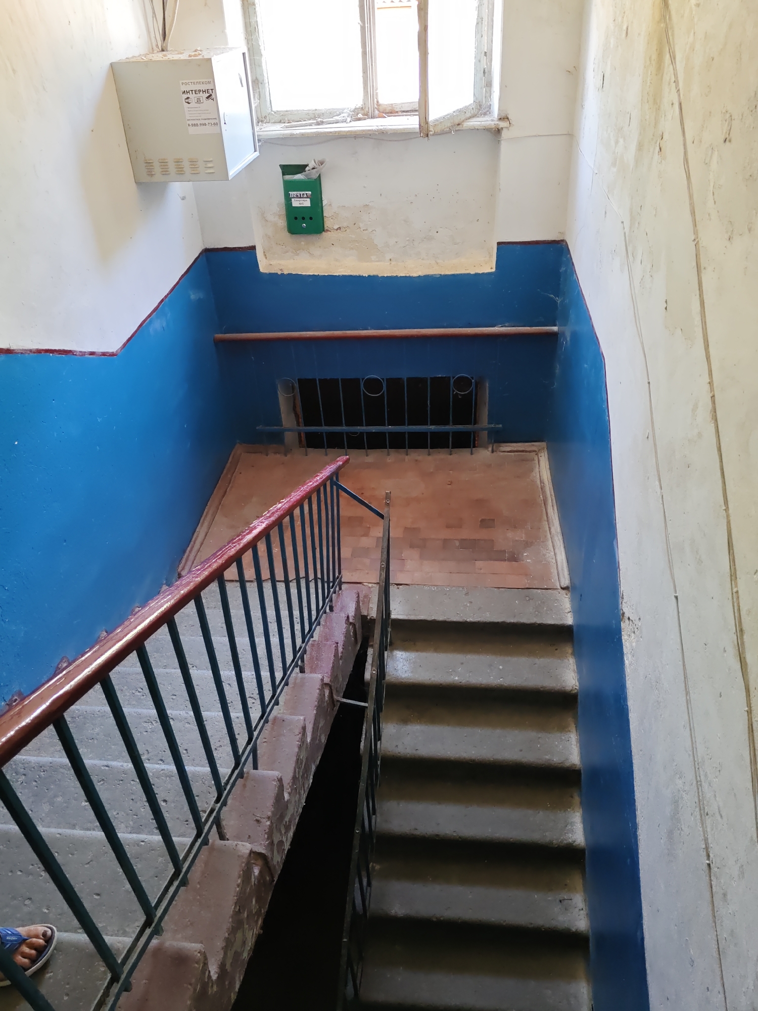 This is an interior image of a dimly lit apartment building stairwell in Nizhnedonskoy, Russia. The walls are painted with a distinctive blue wainscoting. A set of concrete stairs with a dark metal railing and a reddish-brown wooden handrail descends to the right. The lower portion of the stairwell is partially visible and appears darker. At the top of the stairs, a barred opening is visible in the blue wall. To the left of this opening, a green metal mailbox is mounted on the wall, with a white electrical box above it. A window with a wooden frame is situated above the mailbox and electrical box. The floor at the landing area appears to be tiled with reddish-brown bricks. The overall condition of the stairwell suggests it is aged and may be in need of maintenance, with peeling paint and some dust visible. A person's foot in a blue sandal is visible on the bottom left stair.
