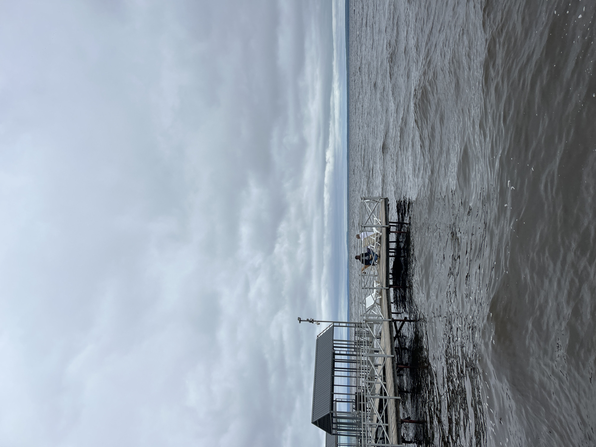 The image depicts a cloudy day at a pier in Kostovaty, Russia. A dark, overcast sky stretches across the frame, with the sea below appearing choppy and dark. The pier, constructed of wood and metal railings, extends into the water. On the pier, a small gazebo-like structure with a dark roof is visible. Two individuals are seated on lounge chairs on the pier, facing away from the camera and towards the water, appearing to be engaged in conversation. The overall atmosphere is somber, with the weather contributing to a sense of muted light and activity.