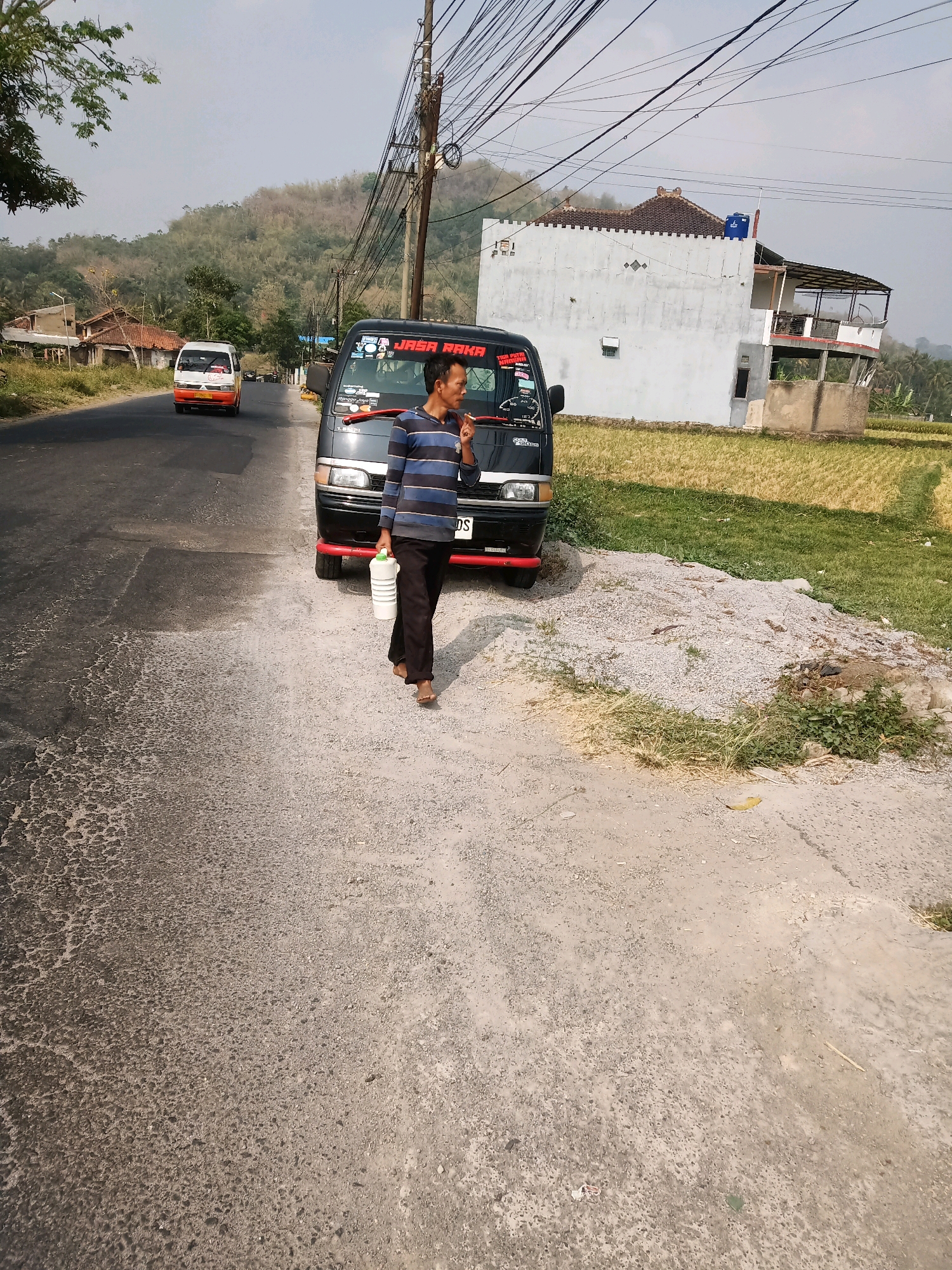 A man in a striped shirt and dark pants walks along a dirt shoulder next to a paved road, carrying a white plastic container. He is barefoot and appears to be smoking a cigarette. A black van with red trim is parked on the shoulder. In the background, a white building with a tiled roof and balconies stands next to a field. A white and orange van is driving away on the road. The scene is set against a backdrop of a grassy hillside and a cloudy sky. The location is near a rural area in Garut, Indonesia.
