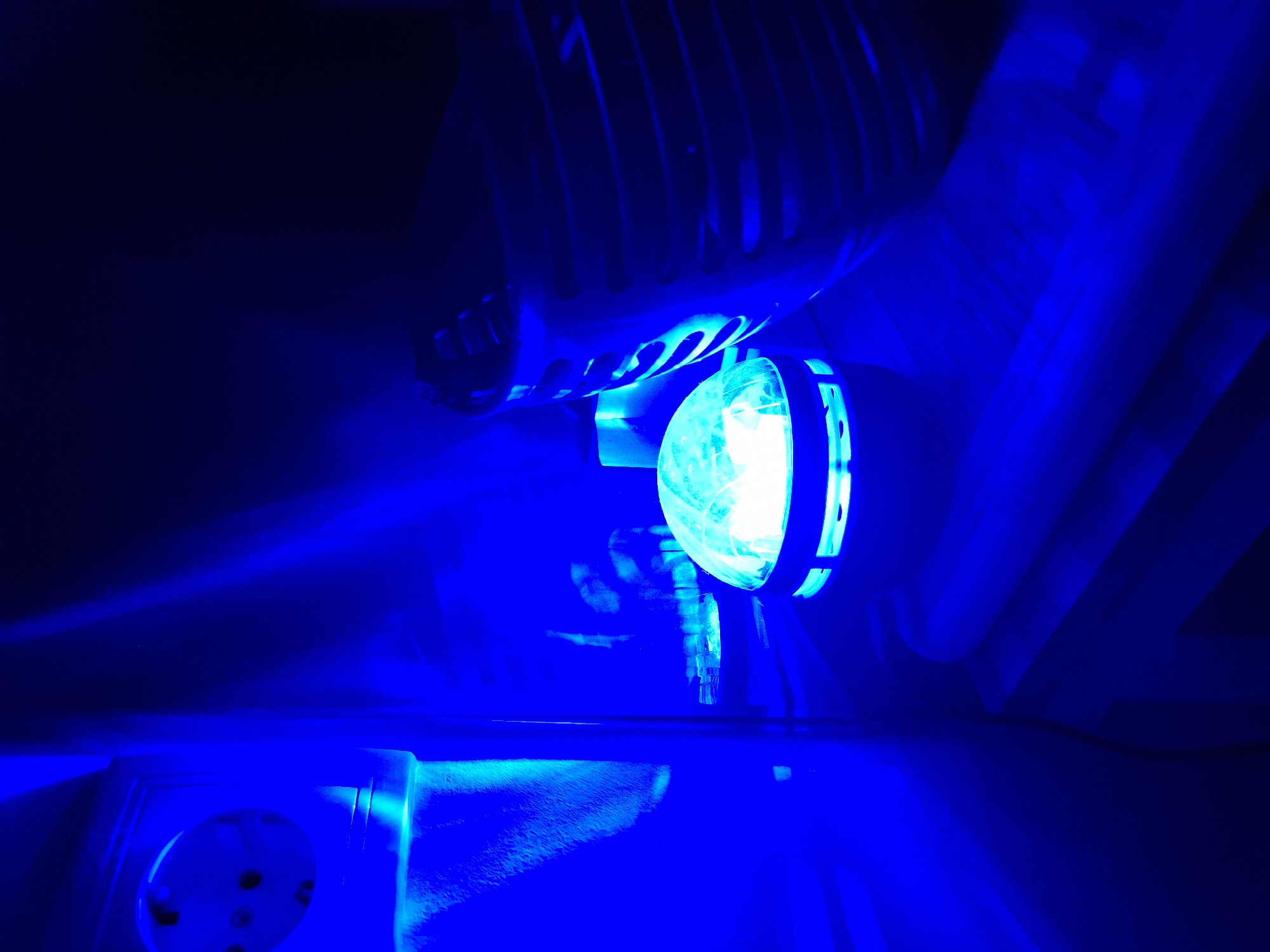 The image displays an indoor scene intensely illuminated by a bright, electric blue light. A prominent circular light fixture, emitting a powerful, almost white-blue glow, is situated in the upper-middle section of the frame. This light source has a dark casing and reveals some internal components through its transparent or translucent cover.
Above and slightly behind the light fixture, a structure resembling a grille, vent, or fan cover with horizontal slats is visible, also bathed in the blue light. In the lower-left corner, a part of a light-colored electrical outlet or socket with multiple circular holes is discernible. The surrounding areas and background are largely dark blue, with subtle variations in tone created by the light and shadow. The surfaces appear indistinct, possibly walls or other equipment within an enclosed space.
No people are present, and no specific activity is taking place; the image is a static capture of the illuminated environment. The dominant blue light creates a monochromatic, high-contrast aesthetic, suggesting a technical, modern, or possibly decorative setting. There are no visible location cues beyond the electrical outlet, which hints at an indoor, artificial environment. No text is visible in the image.