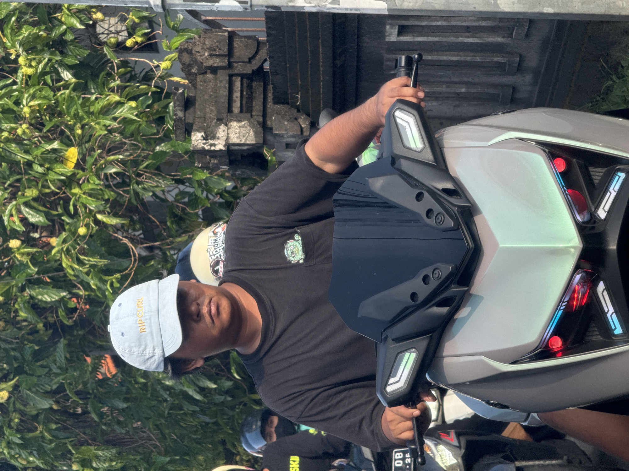 The image captures a man riding a motorcycle in North Kuta, Indonesia. He is wearing a black long-sleeved shirt, a light blue "RIP CURL" baseball cap, and a helmet underneath. The motorcycle is silver with a black front panel. The scene is set outdoors with lush green foliage visible in the background. A stone structure, possibly a building facade or entrance, can be seen behind the rider. Other people on motorcycles are present in the background.