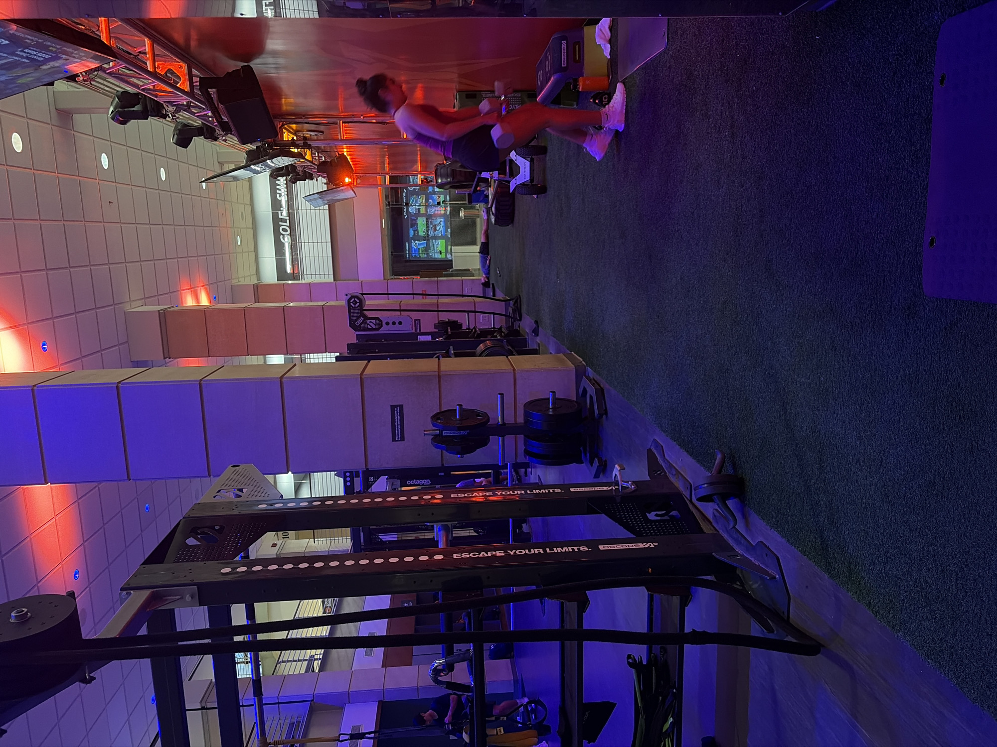 The image shows the interior of a gym located in Austin, Texas. A woman is exercising with weights, lying on her back with her legs bent and feet on a platform, lifting the weights as part of her workout. Gym equipment, including a squat rack, is visible. The gym has artificial turf and is illuminated by a combination of red and blue lighting. Signage such as "GOLF SIM" and "ESCAPE YOUR LIMITS" are visible on the equipment. The interior design includes a tiled wall in the background.