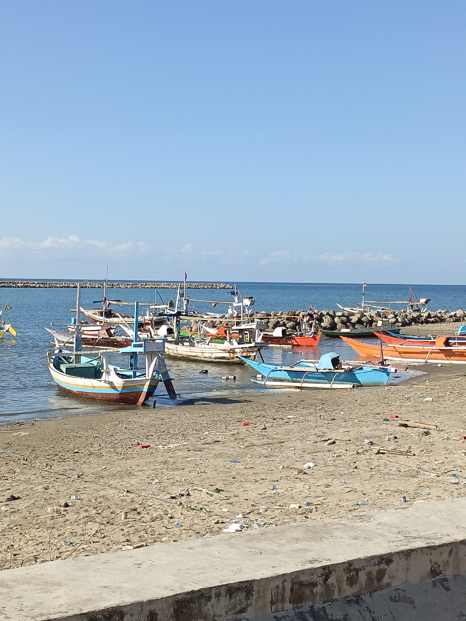 This is a medium-shot, eye-level, vertical, outdoor, daytime, photograph of a beach in Ela-ela, Indonesia, littered with various forms of trash. The beach is composed of light brown sand. In the foreground, a concrete barrier runs horizontally across the bottom of the frame. Beyond the barrier, the sand is covered with debris including plastic bottles, wrappers, and wooden scraps. Several small, colorful fishing boats are docked in the shallow water close to the shore. One boat, predominantly white with blue and yellow stripes along its hull, is pulled up onto the sand. Further out in the water, more fishing boats of various sizes and colors are anchored or tethered. The water is a clear blue, and beyond the boats, a breakwater made of large rocks extends across the horizon. The sky is a clear, bright blue with a few wispy white clouds. There are no people visible in the frame.