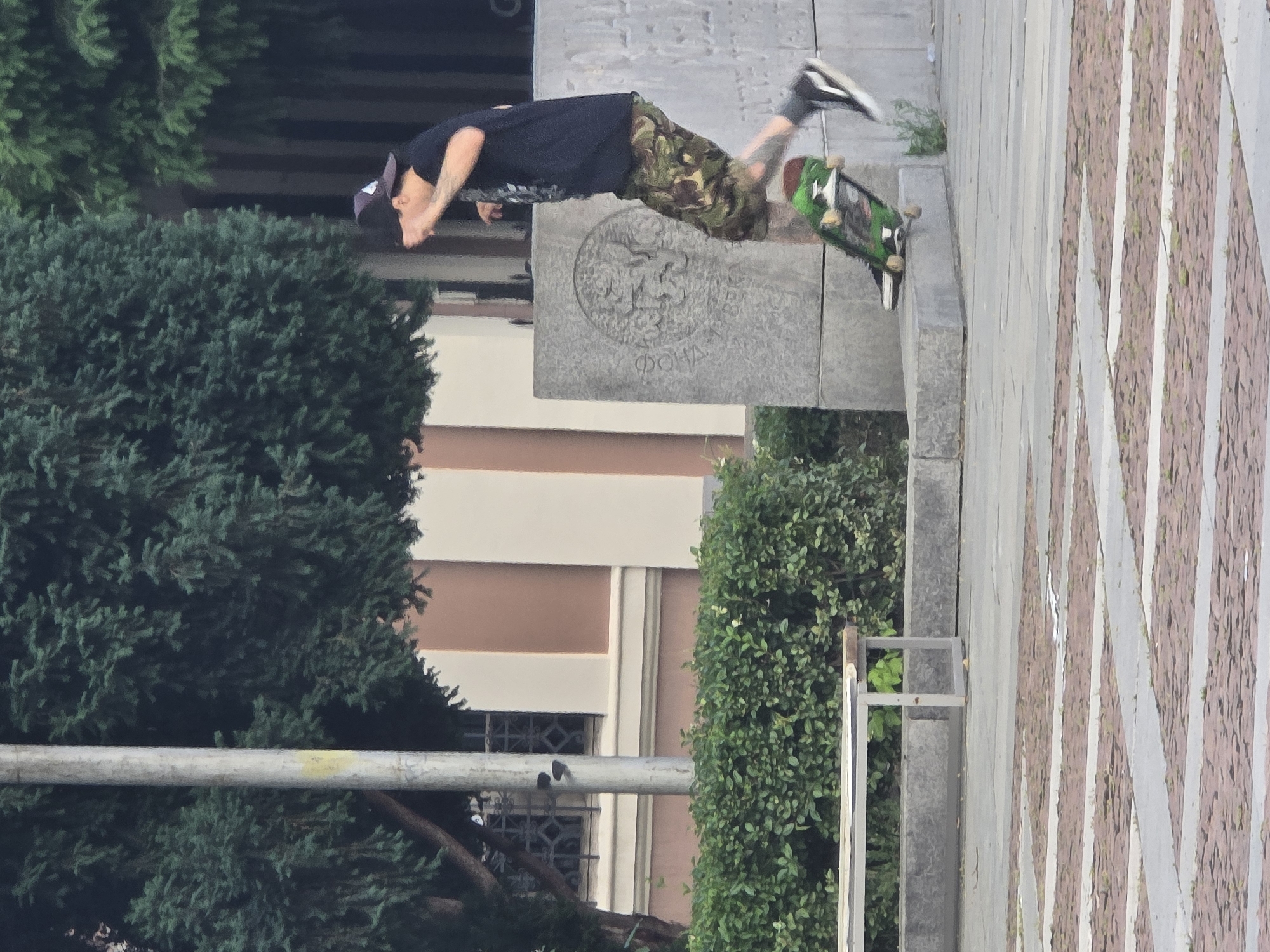 A young man is performing a skateboarding trick outdoors in a public square in Plovdiv, Bulgaria. He is mid-air, with his skateboard beneath him, as if jumping off a ledge. The man is wearing a dark t-shirt, camouflage shorts, a baseball cap, and sneakers. He has tattoos on his arm. In the background, there are large evergreen trees, a building with beige and brown sections, a stone monument with an engraved emblem, a manicured hedge, and a park bench. The ground is paved with light and dark square tiles arranged in a diamond pattern. The lighting suggests it is daytime.