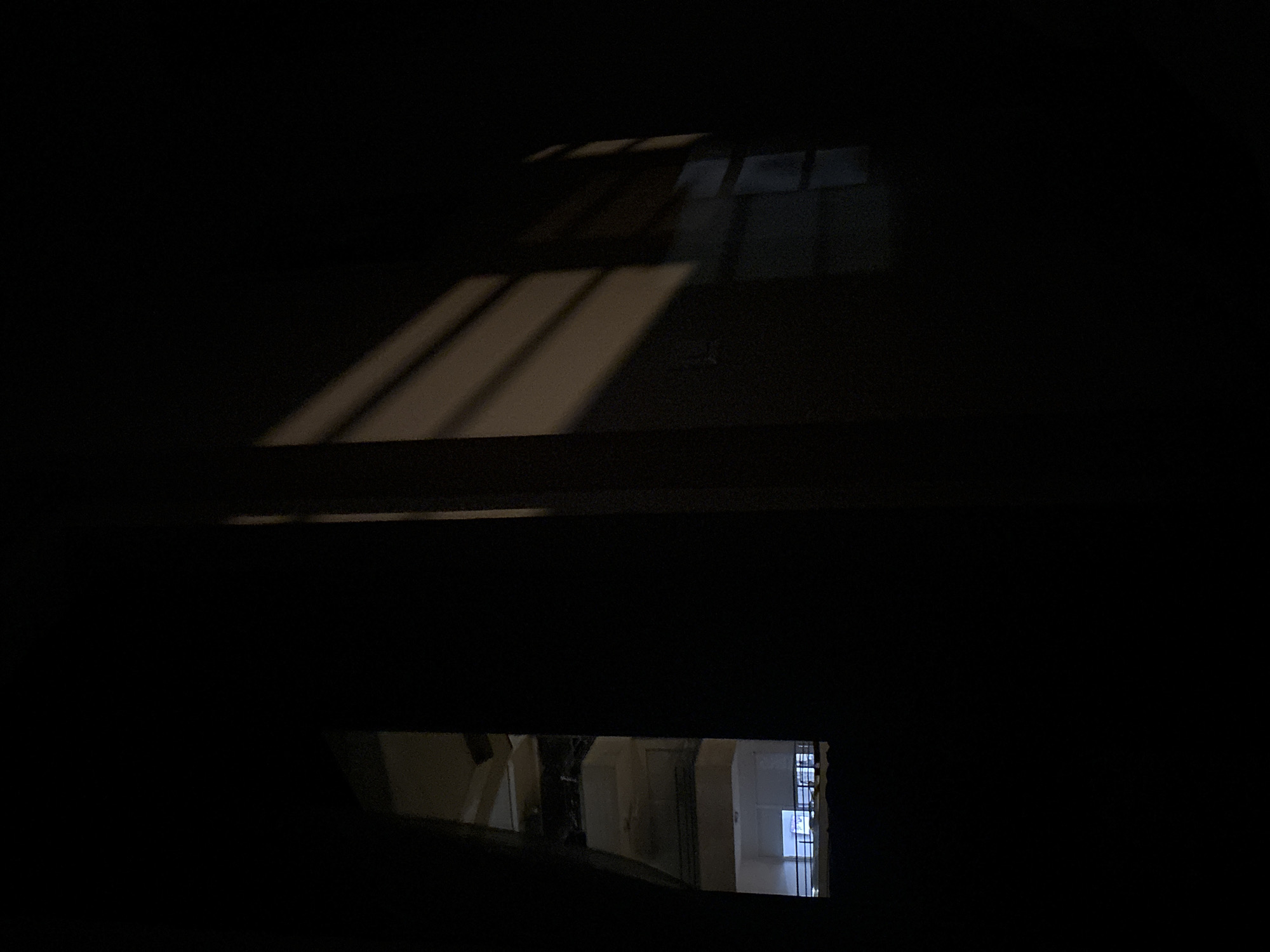 A dark, indoor scene with light patterns cast from a window above, illuminating parts of the wall. In the lower portion of the image, a rectangular opening reveals a view of an interior space with light walls, doors, and a barred window. The location is unknown and cannot be determined.