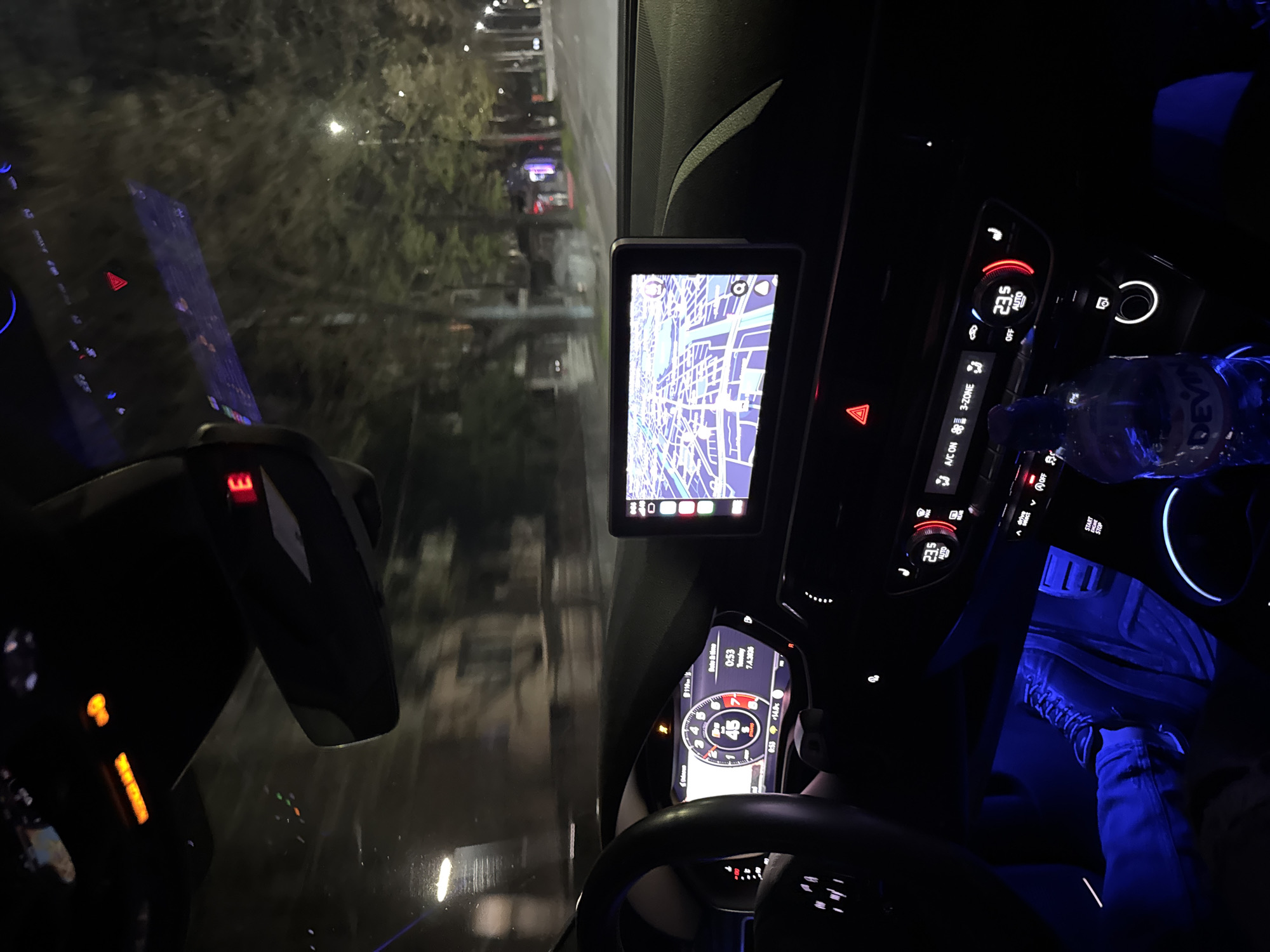 The image shows the interior of a car at night. The dashboard displays a map on a screen and a speedometer showing 45 km/h. A bottle of water is visible next to the climate control panel. Blue lighting illuminates the footwell, revealing a person wearing jeans and sneakers. The view through the windshield shows streetlights and trees, indicating an urban setting. Without more information, determining a specific location in Plovdiv, Bulgaria, is not possible.