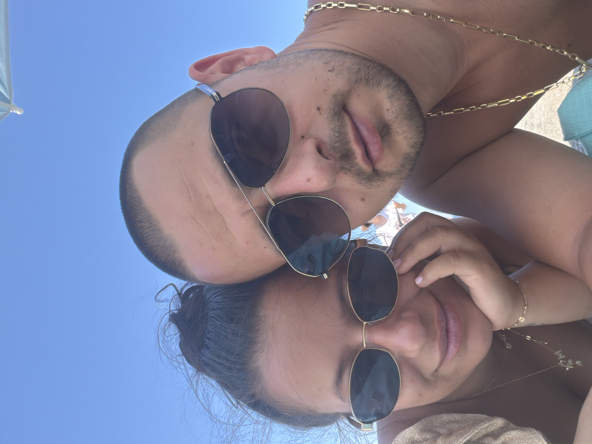 A close-up, eye-level, selfie-style photograph shows a young couple posing against a clear blue sky. The woman is on the left, and the man is on the right, with their heads angled towards the camera. Both are wearing sunglasses. The woman has dark hair pulled up, and the man has a shaved head and a light beard. The man is wearing a gold chain. They appear to be at a beach, as indicated by the sand visible in the lower portion of the frame and a light blue beach umbrella in the upper right corner. The lighting suggests it is daytime. There are no specific landmarks visible that would indicate the location within Atiya, Bulgaria.