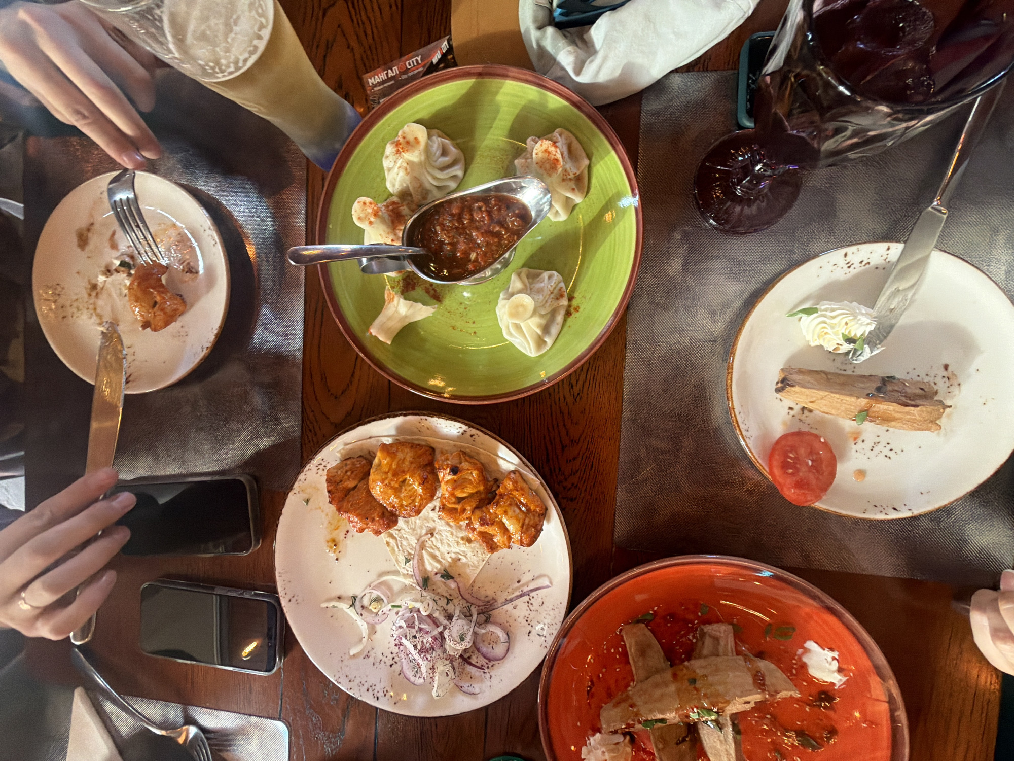 An overhead shot captures a dining table set with multiple dishes and drinks, indicating an ongoing meal. The table is dark wood, partially covered by dark, textured placemats.
Several hands are visible, holding cutlery or resting near items. On the left, a hand holds a fork over a white plate with remnants of a partially eaten dish, possibly grilled chicken or meat. Another hand holds a knife. Two smartphones with dark screens are also on the table.
Central to the image is a vibrant green plate featuring five khinkali (Georgian dumplings), some lightly dusted with spices, arranged around a metal serving spoon filled with a rich, dark red sauce. Below this, another white plate holds several pieces of grilled meat, likely chicken, served on a flatbread (possibly lavash) and garnished with finely sliced red onions and herbs.
To the right, a white plate presents a dessert or pastry, shaped like a log, accompanied by a dollop of whipped cream and a slice of red tomato, which might be a garnish or part of a savory course. Below this, a red plate contains another dish, possibly rolls or appetizers, topped with a white sauce or cream and sesame seeds.
Drinks include a tall glass of light-colored beer with foam and a dark glass holding a red beverage, possibly wine or a berry drink, with ice cubes. The lighting is warm and somewhat dim, suggesting an evening meal in a cozy restaurant or cafe setting. A small card with the text "МАНГАЛ CITY" is visible near the green plate, indicating the establishment's name.