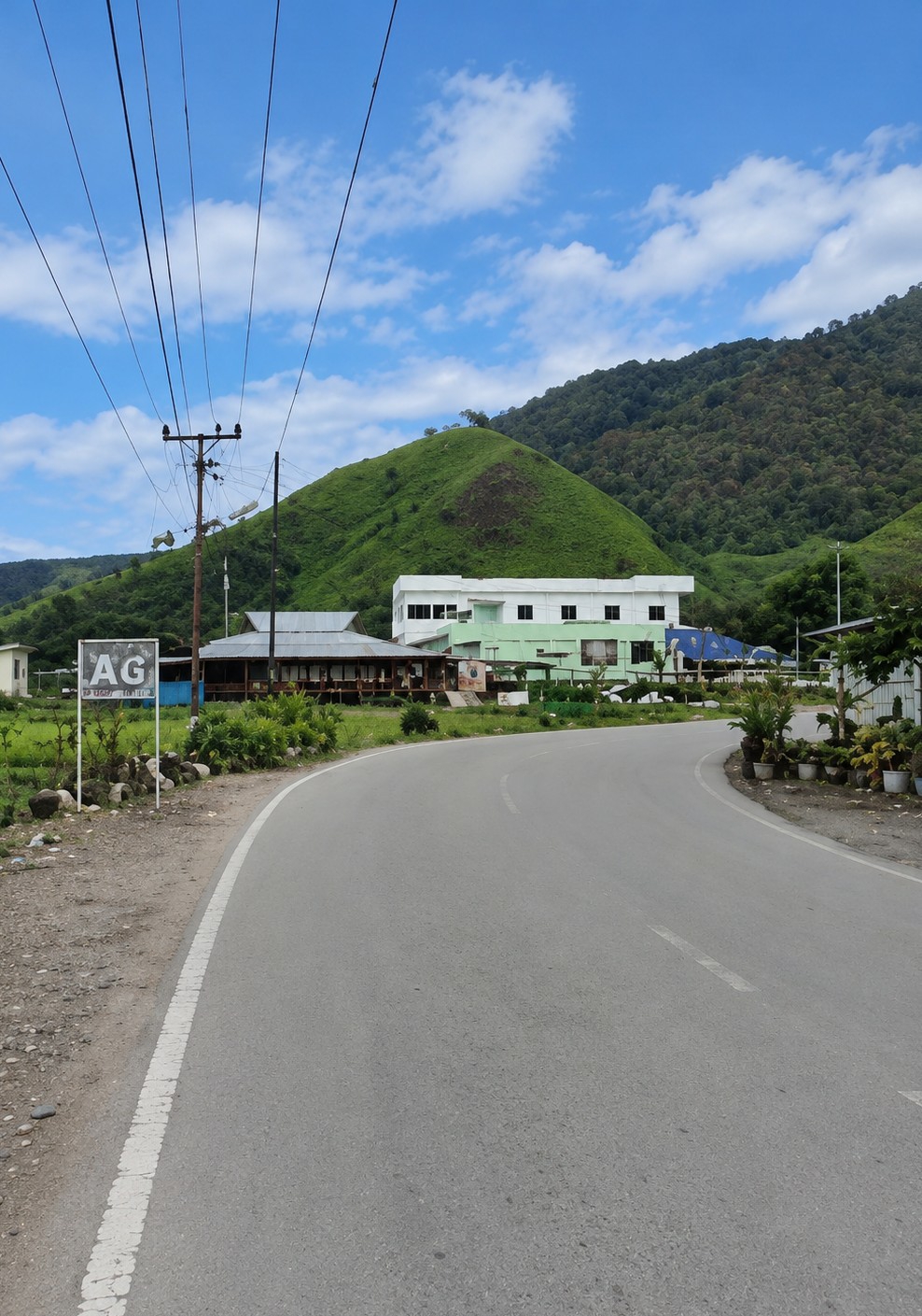 The image shows a road curving through a landscape dominated by lush green hills under a partly cloudy blue sky. On the left side of the road, there is a sign with the letters "AG" in large white block letters. Behind the sign, there are telephone poles with numerous wires stretching across the frame. Further back, a low-lying building with a corrugated metal roof is visible, and beyond that, a larger, multi-story building with a white upper section and a light green lower section. This building appears to be modern with many windows. The surrounding environment is verdant, with trees and foliage visible on the hillsides and closer to the buildings. The road is paved and has white lane markings. There are some potted plants and what appear to be stones or small rocks along the side of the road near the buildings. The overall scene suggests a rural or semi-rural setting within a mountainous region.