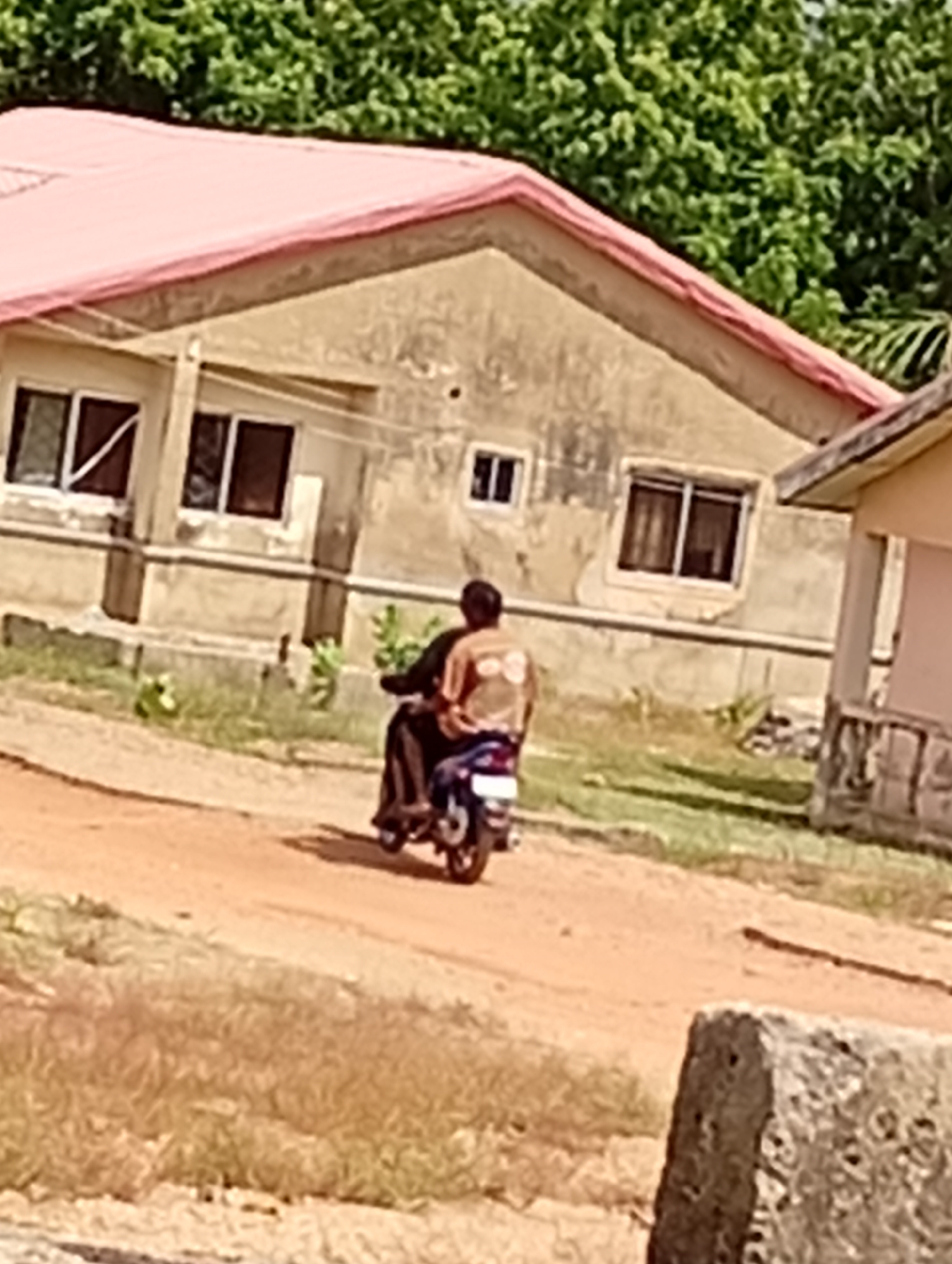 The image depicts a man riding a motorcycle on a dirt road in a suburban neighborhood in Unknown, Unknown. The man has dark hair, is wearing a brown shirt, and is carrying a passenger. The motorcycle is blue. In the background, there is a tan one-story house with a red roof and four windows, and some trees. In the foreground, there is a dirt road and some grass. There is also a stone object that is partially visible in the lower right corner of the image.