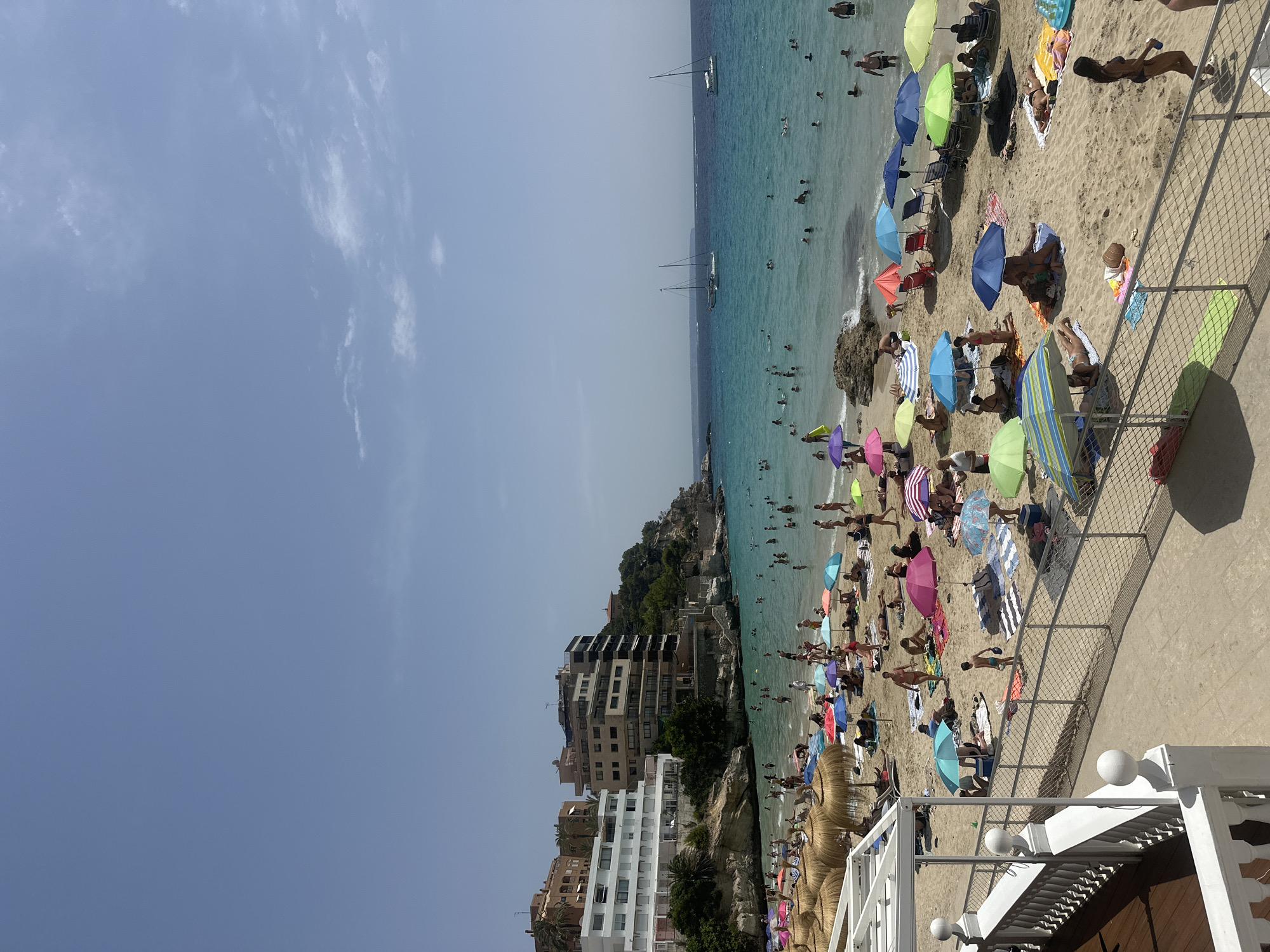 A high-angle, full shot captures a bustling beach on a sunny day in Sant Agustí, Spain. The foreground features a white railing and wooden deck, offering a vantage point over the sandy shore. The beach is densely populated with beachgoers of all ages, some sunbathing on towels and loungers, others wading and swimming in the clear, turquoise waters. A multitude of colorful umbrellas, both straw and fabric, dot the sand, providing shade.
In the mid-ground, buildings line the cliffside overlooking the beach, with a mix of white, beige, and brown structures, some appearing to be hotels or apartment complexes. Lush green trees and foliage cling to the rocky terrain. Several sailboats are visible in the distance, anchored in the calm sea, with a faint landmass on the horizon. The sky is a clear, pale blue with a few wispy white clouds. The overall atmosphere is one of a lively, popular tourist destination.