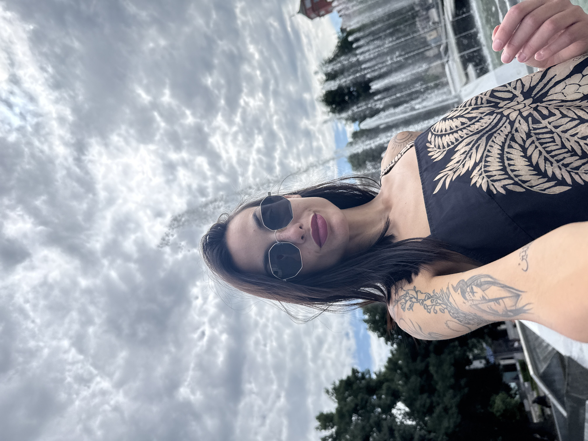 The image is oriented on its side, depicting a young woman posing for a selfie outdoors in what appears to be a public park or square in Pleven, Bulgaria. She has long, dark hair, is wearing dark sunglasses with geometric frames, and has dark red lipstick. Her attire includes a dark-colored sleeveless top or dress featuring a prominent light-colored, intricate patterned design resembling leaves or tribal motifs on the chest.
Several tattoos are visible on her arms. Her left arm displays a large, elaborate tattoo that seems to depict a mythical creature or bird intertwined with flowing, possibly floral, elements. On her right arm, a smaller, circular tattoo is visible, which includes what appears to be an "Om" symbol. She is looking directly towards the camera with a neutral to slightly smiling expression.
The background is dominated by a heavily clouded sky, featuring textured grey and white stratocumulus clouds. Behind her, multiple jets of water from a fountain create blurred vertical streaks due to motion, contributing to the dynamic feel of the scene. Dark green trees are visible beyond the fountains, and a distant reddish building can be faintly seen above the tree line, suggesting an urban setting. The overall weather appears overcast during daytime.