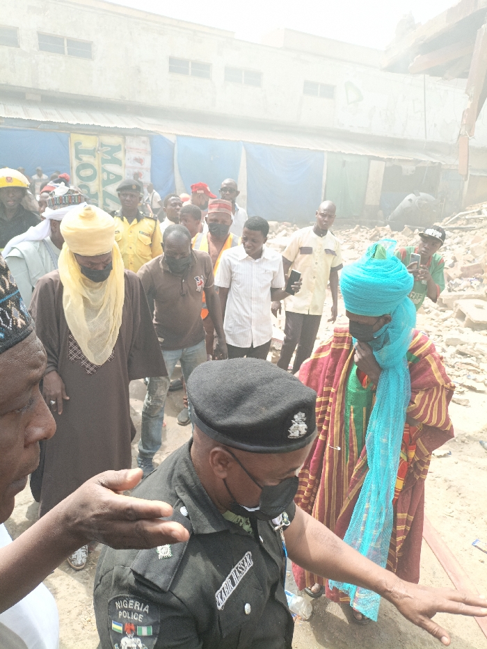The image depicts an outdoor scene in Fagge C, Nigeria, characterized by a crowd of individuals amidst rubble and debris. The ground consists of loose dirt and scattered construction material fragments. In the background, a light-colored building, possibly damaged or undergoing demolition, is visible, partially obscured by dust or haze. Blue tarpaulins or temporary structures are situated below this building.
In the immediate foreground left, an adult male, with dark skin, wears a patterned cap and a light-colored long-sleeved garment. He extends his right hand with an open palm, gesturing towards the center. Adjacent to him, a Nigerian Police officer, identifiable by his black beret with an emblem, dark uniform bearing "NIGERIA POLICE" and "KABIR HASSAN" on a nametag, and shoulder insignia, looks downwards and slightly to his right. He wears a black face mask. To the officer's right, a person in traditional attire, including a red and yellow striped robe and a bright turquoise head wrap that covers the lower face, is observed.
In the midground, numerous individuals are present. Notable figures include a person in traditional yellow attire with a turban and face mask, another in a brown short-sleeved shirt and black face mask, and a male holding a mobile phone horizontally as if recording or photographing. A man in a green t-shirt and black baseball cap is also visible holding a phone in this manner. The crowd generally stands and observes the scene, with some individuals looking towards the foreground figures.